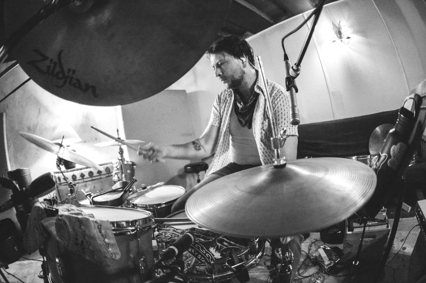 A man playing the drums in a recording studio with microphones set up around him, black and white photo.