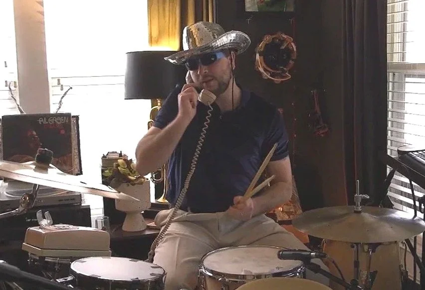 Man wearing a large, shiny cowboy hat and sunglasses, sitting at a drum set in a room, talking on a landline phone, holding drumsticks, with a vinyl record on a turntable nearby.