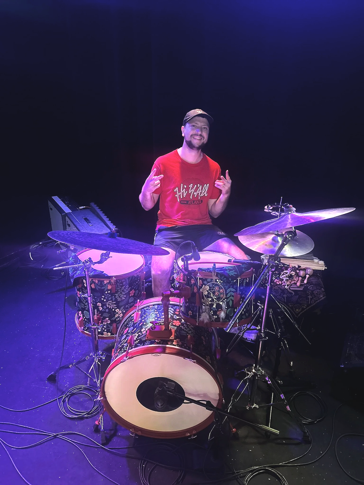 A man in a red t-shirt and black cap sitting behind a colorful drum set on a dark stage, smiling and making a peace sign with both hands.
