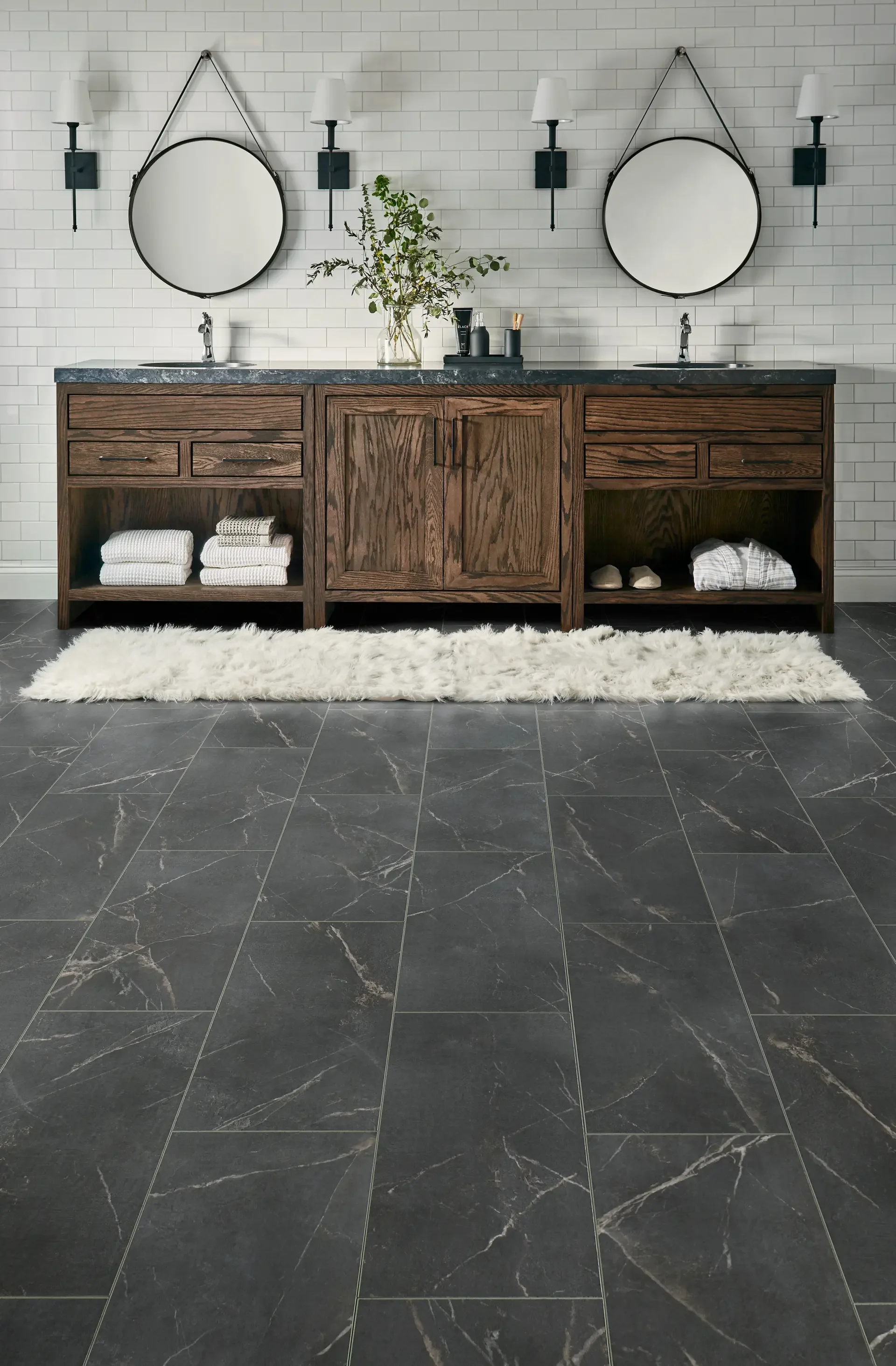 Bathroom vanity with two sinks, round mirrors, wall-mounted lamps, dark marble countertop, wooden cabinet, white towels, green plant, soap dispensers, and a white rug on gray tile floor.