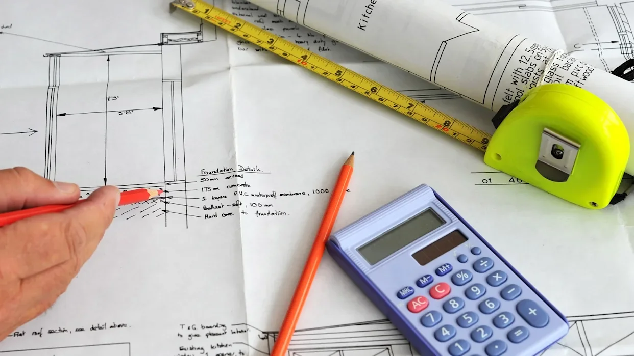 Architectural blueprint with foundation details, a yellow measuring tape, an orange pencil, a calculator, and a rolled-up blueprint on a drafting table.
