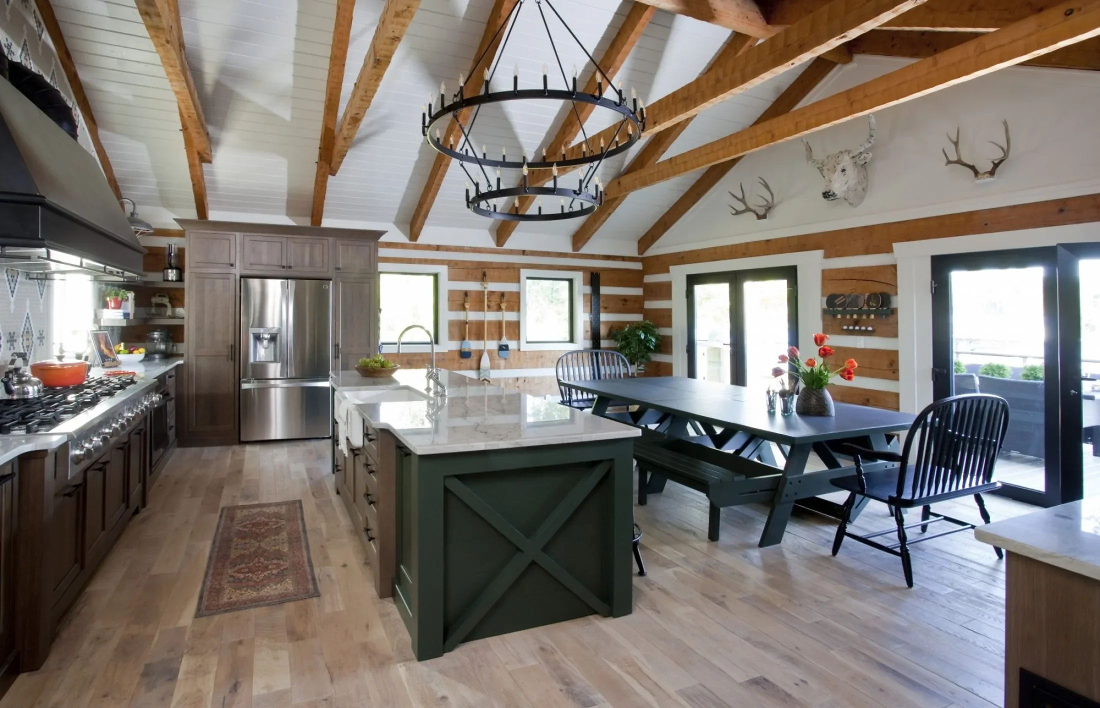 A spacious kitchen with wooden walls and ceiling beams, featuring a large island with a marble countertop, a stainless steel refrigerator, a gas stove, a dining table with benches and chairs, and decor such as antler and deer head wall mounts.