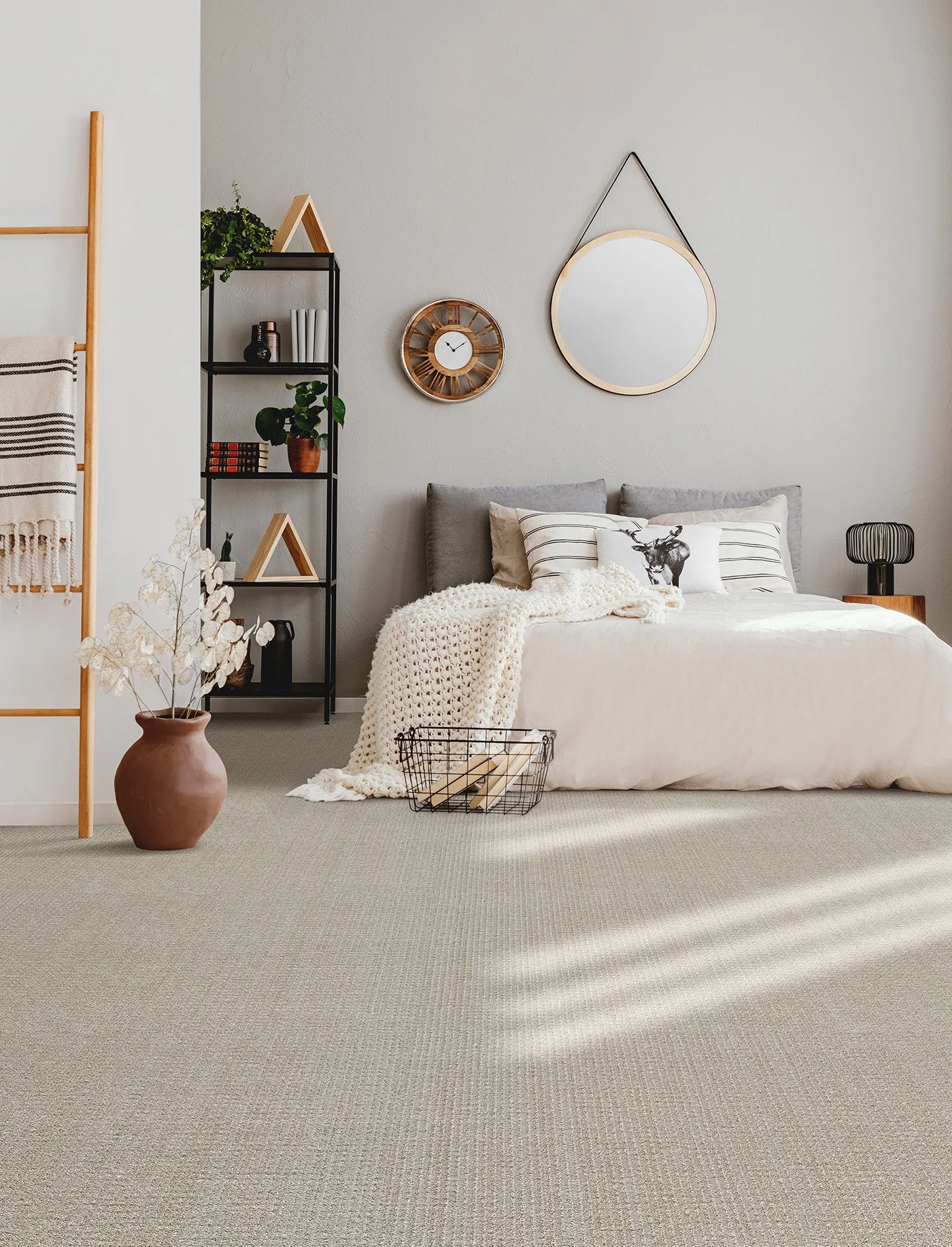 A cozy bedroom with a bed, decorative pillows, a knitted throw, and a wire basket. Decorative wall mirrors and a clock hang above the bed. A black shelving unit with plants and decor items is to the left. A large terracotta vase with dried flowers is on the floor, and a black side table with a lamp is beside the bed.