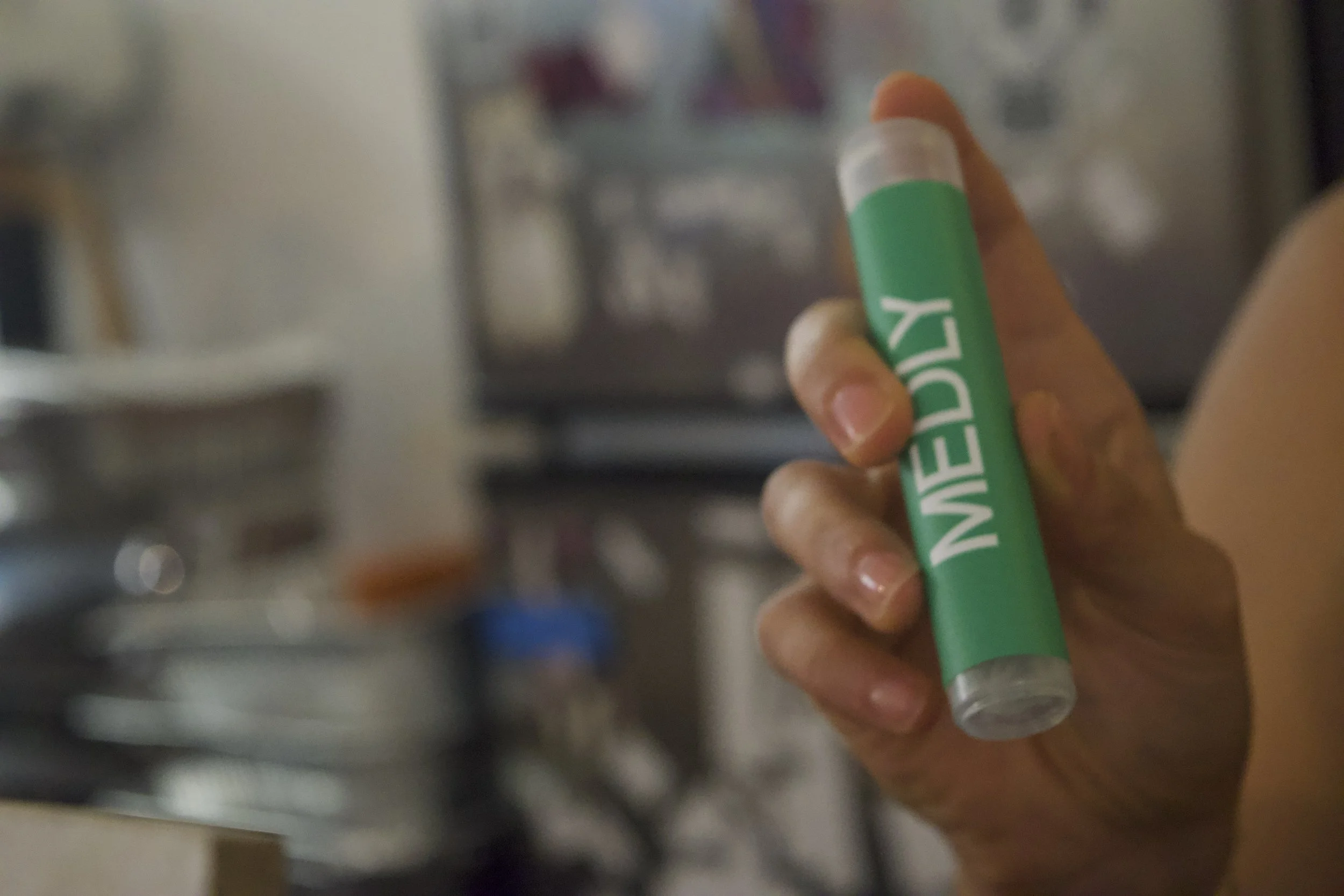 A hand holding a green and white lipstick-style tube labeled 'MEDY' in a cluttered indoor environment.