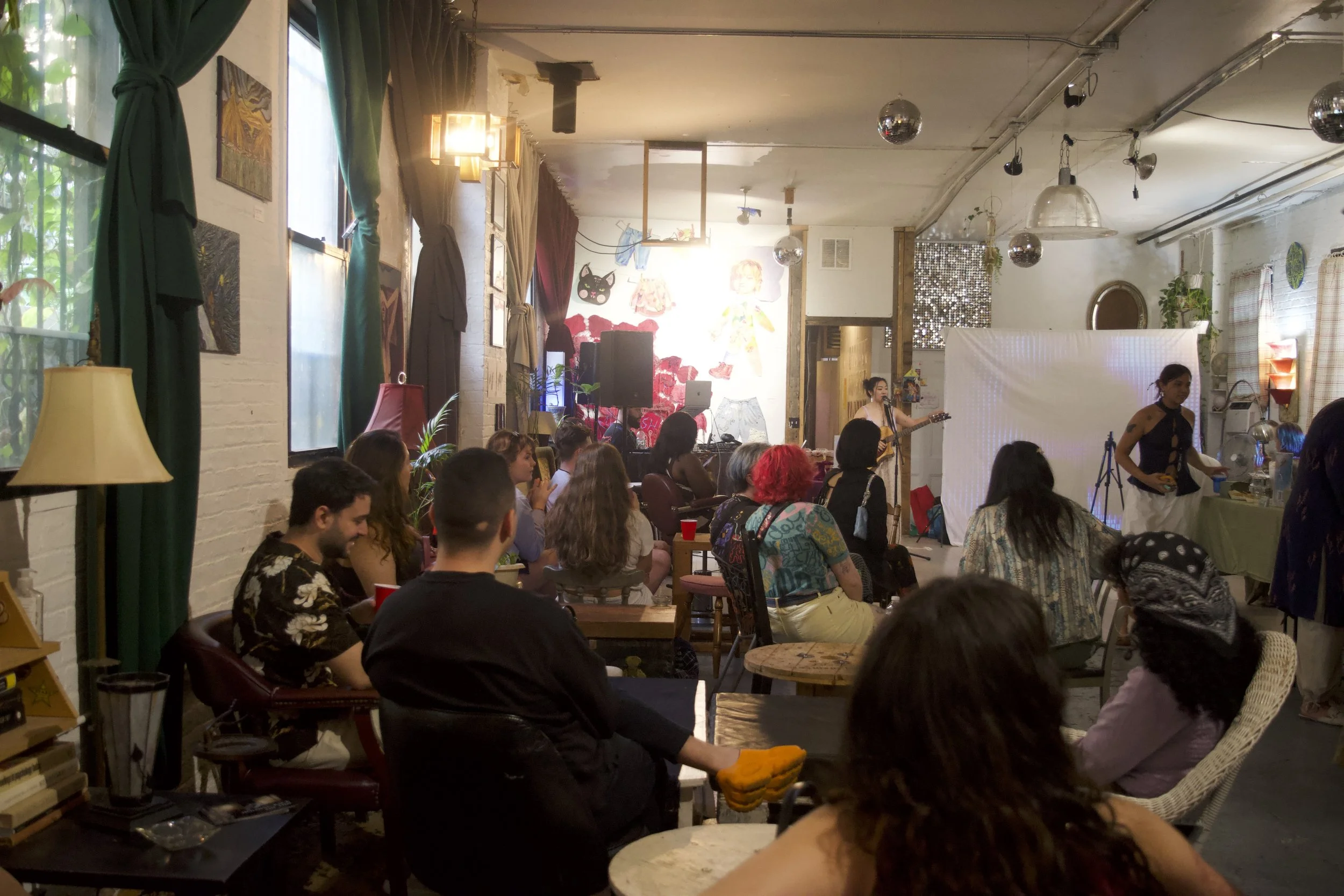 People attending a live music performance in an intimate, cozy indoor venue with colorful decor, artwork on the walls, and a singer playing guitar on stage.