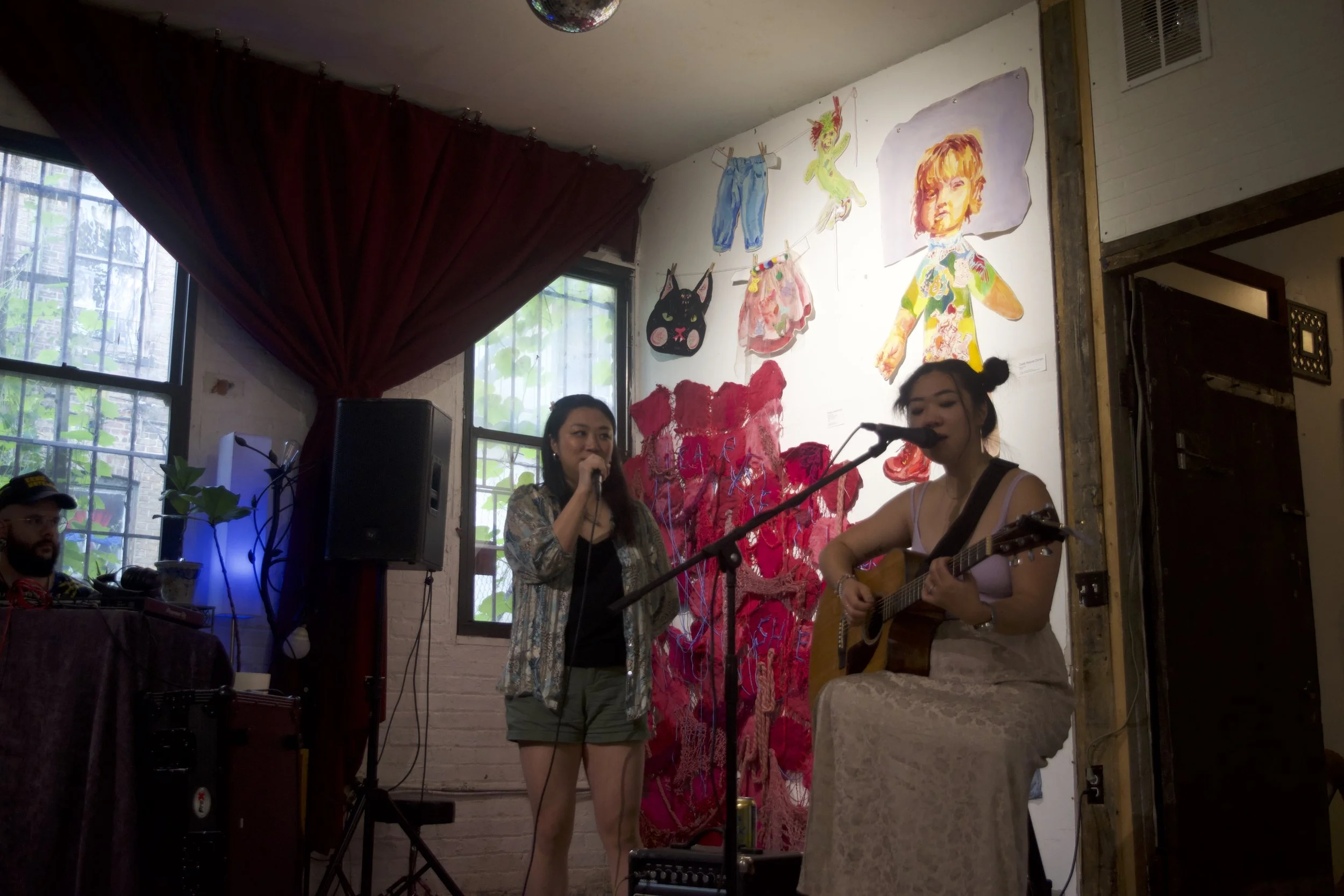 Two women performing music and singing with a microphone and guitar in a cozy indoor setting, decorated with colorful art on the wall, including a large child's drawing, a cat face, and cutouts of clothes and a dragon.