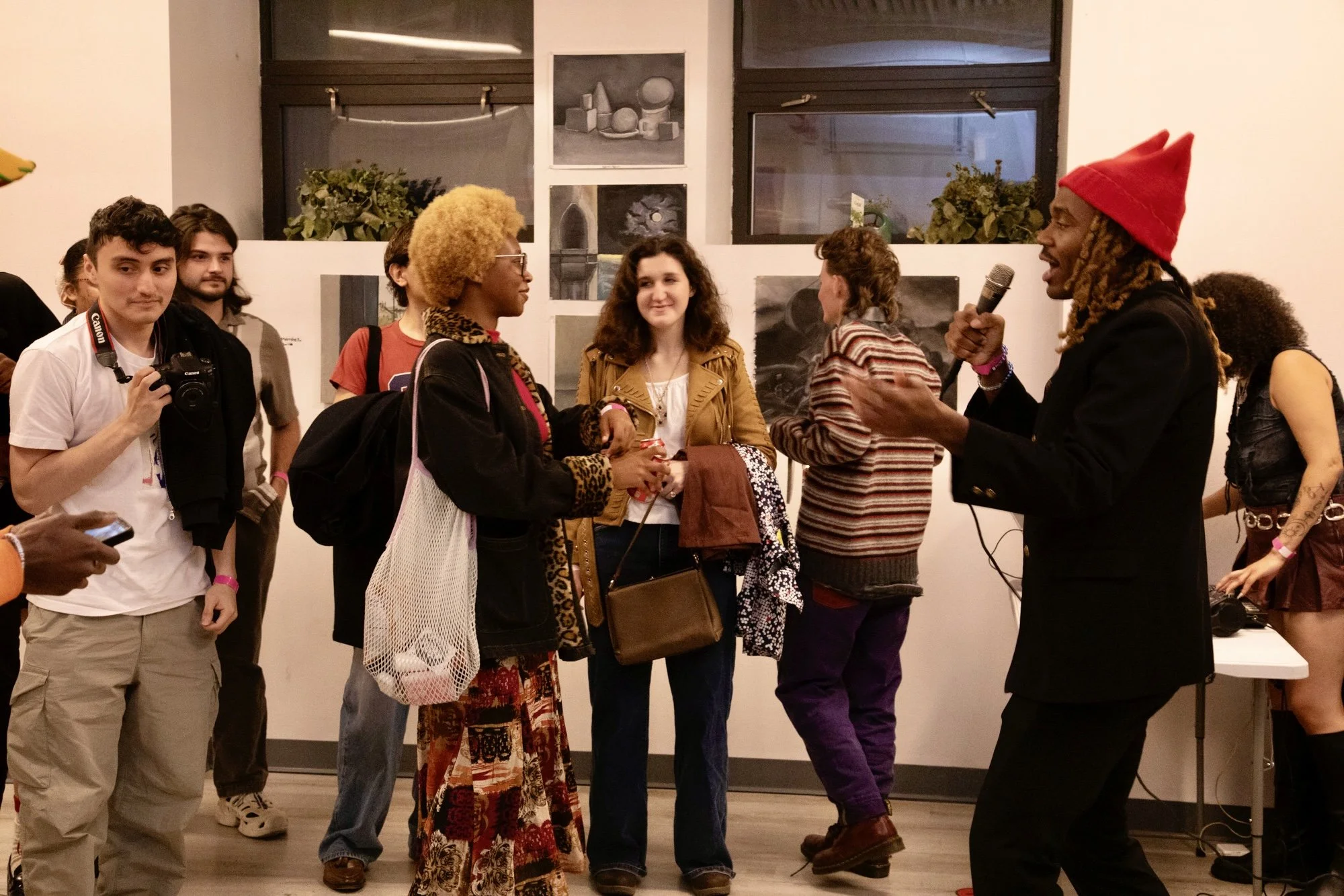 An art gallery event with a man in a black suit and red hat speaking into a microphone, engaging with an audience of diverse young adults. The audience members are listening attentively, some holding cameras or phones, in front of artwork displayed o