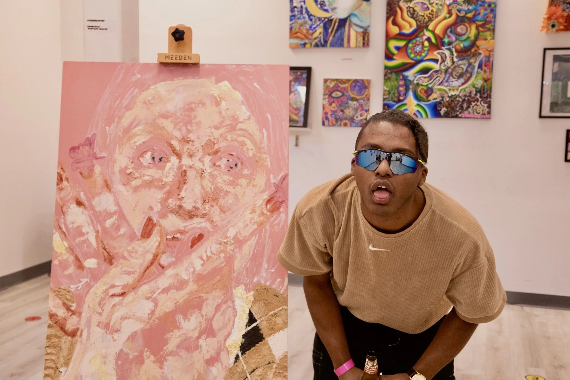 Person wearing sunglasses and a tan Nike shirt leaning forward in art gallery with surreal portraits and colorful abstract paintings in background, next to a large pink portrait of a face with exaggerated features.