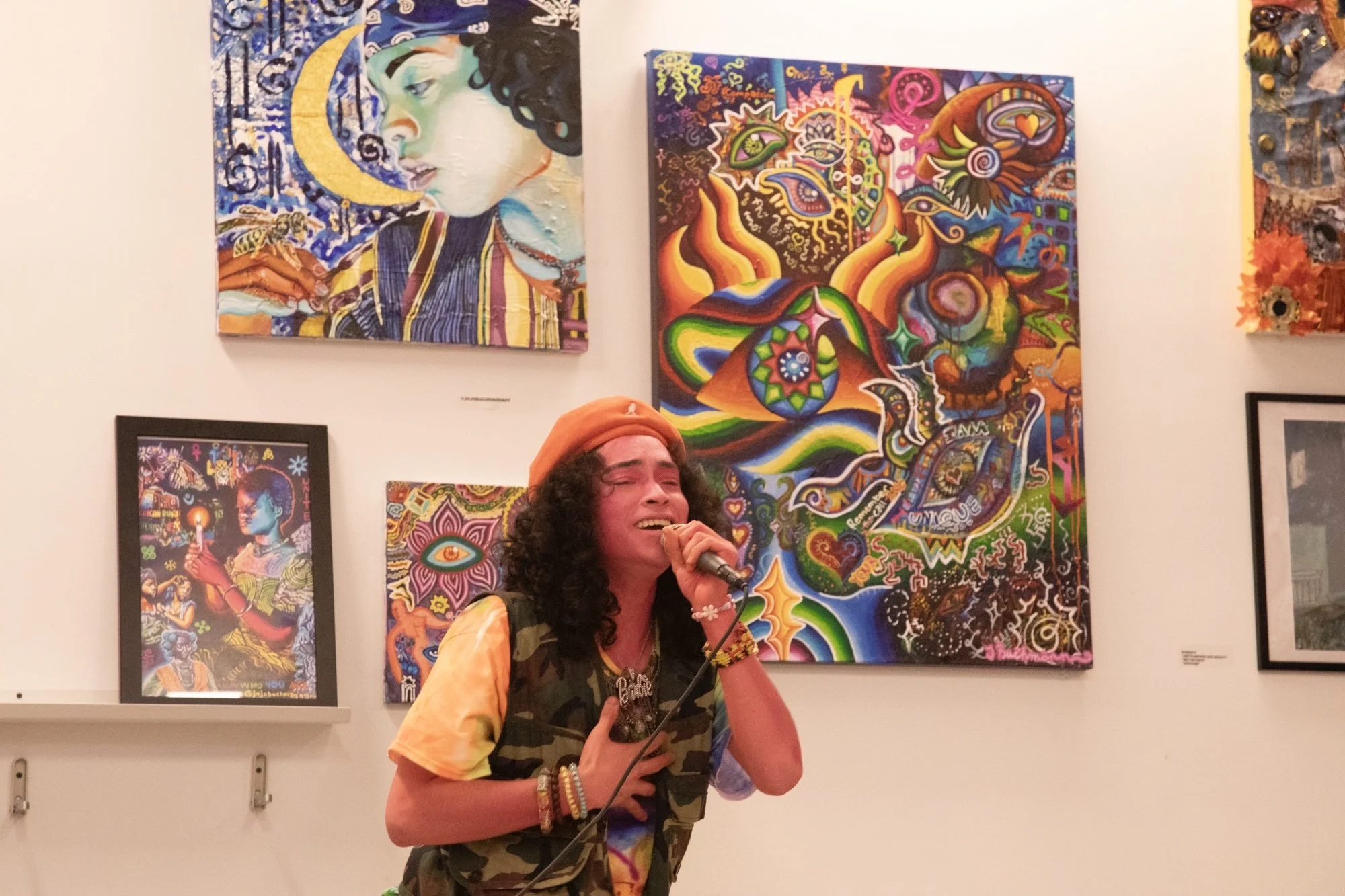 A woman singing into a microphone in an art gallery with colorful abstract paintings on the wall behind her.