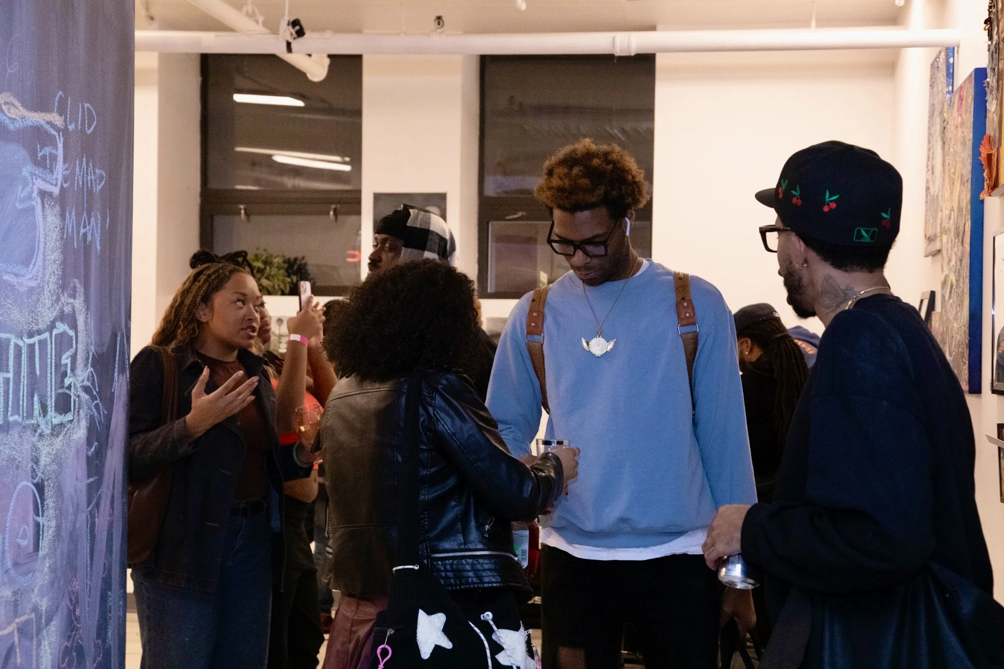 A group of young people socializing at an art gallery, with some engaged in conversation and others taking photos.