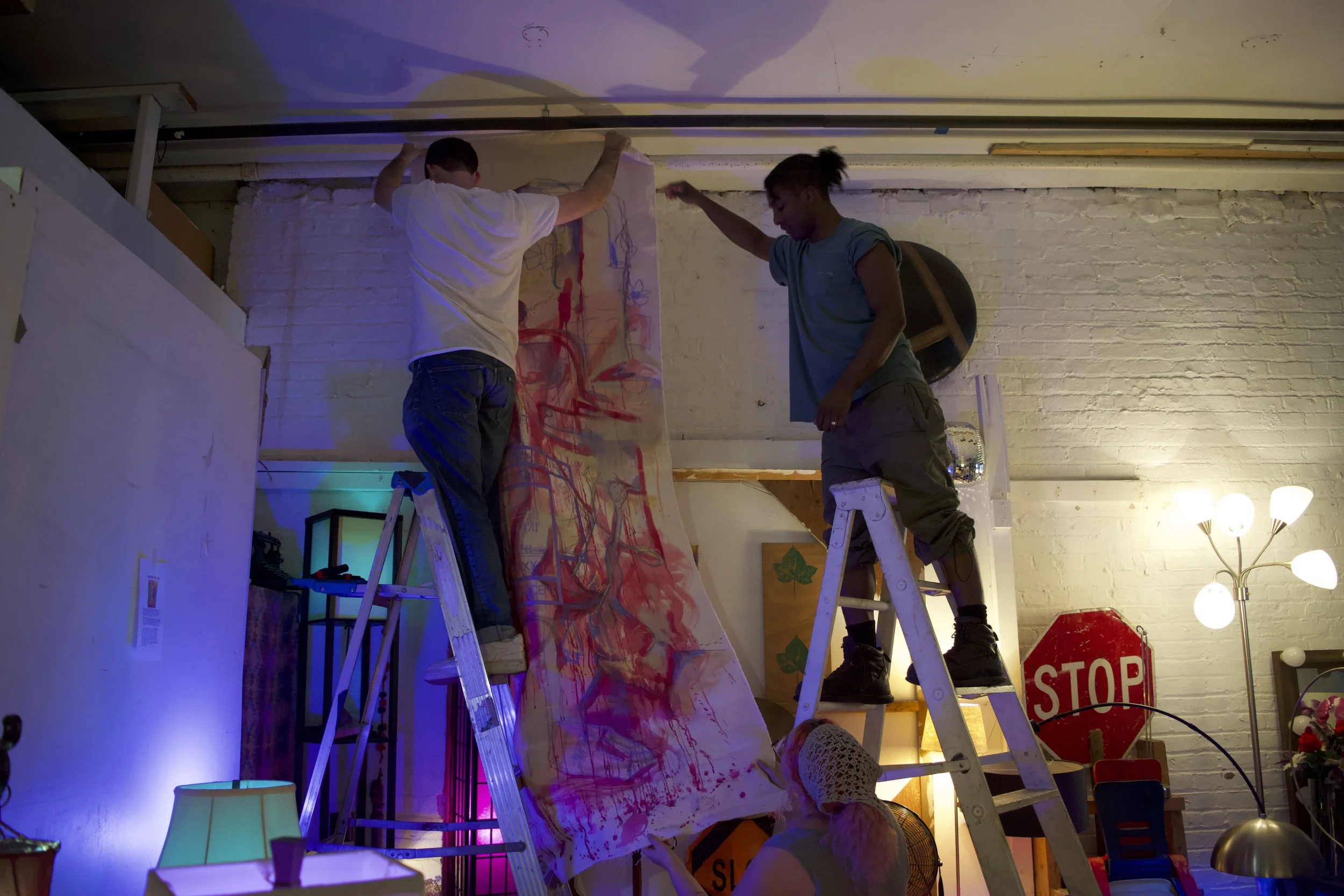 Two people standing on ladders, applying a painted canvas on a wall inside a room decorated with various lamps, artwork, and a stop sign.