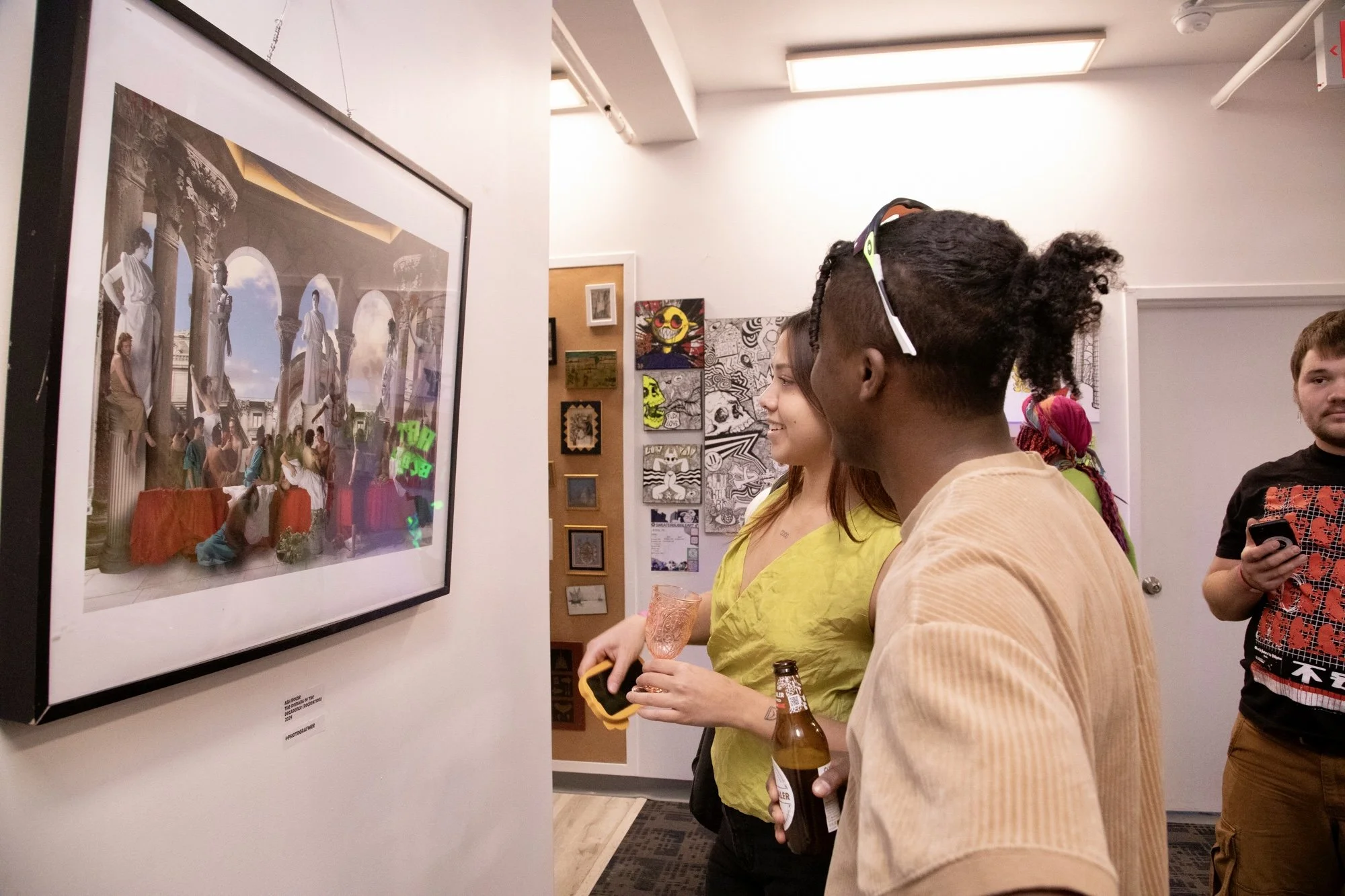 Group of people viewing art in an art gallery, with one person holding a beer bottle and another holding a glass, looking at a framed artwork on the wall.