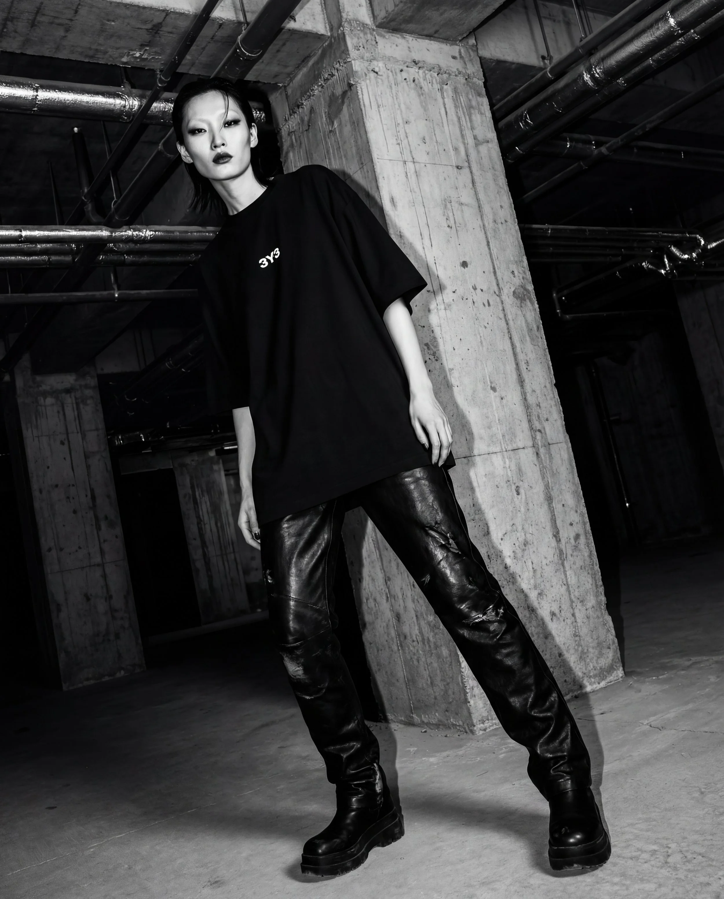 A woman with dark hair and makeup, wearing an oversized black T-shirt Techno Studios by 3Y3, leather pants, and chunky boots, standing against a concrete pillar in an industrial underground parking garage.