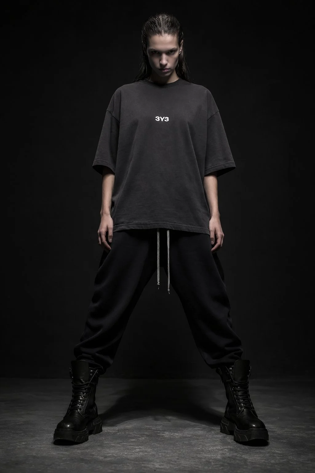 A person standing confidently against a black background wearing an oversized black T-shirt Techno Studios by 3Y3, loose black pants, and combat boots.