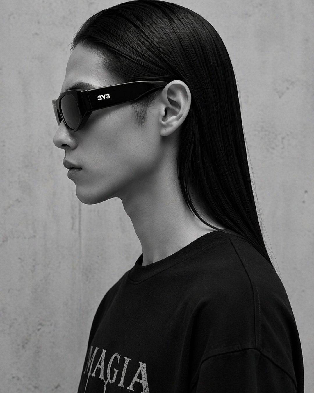 A woman with slicked-back hair wearing large black Techno Studios sunglasses by 3Y3 with white text on the side, facing left against a textured wall.