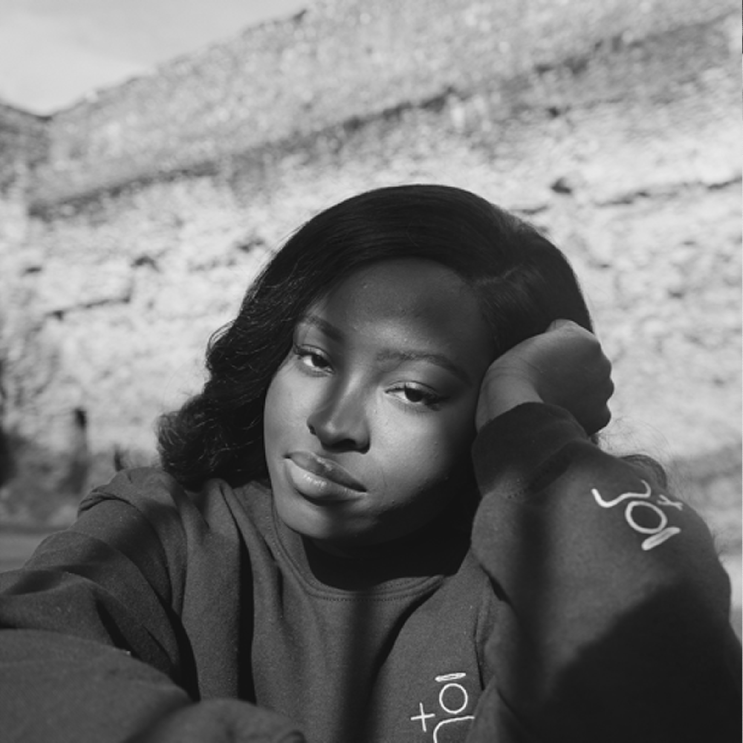 Black and white photo of a woman with wavy hair, resting her head on her hand, in front of a brick wall.