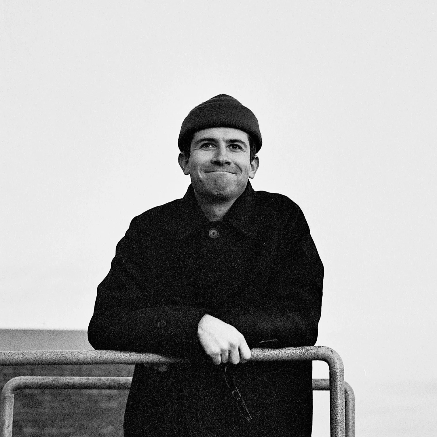 A man wearing a beanie and dark coat, standing behind a railing, smiling and looking at the camera in black-and-white.