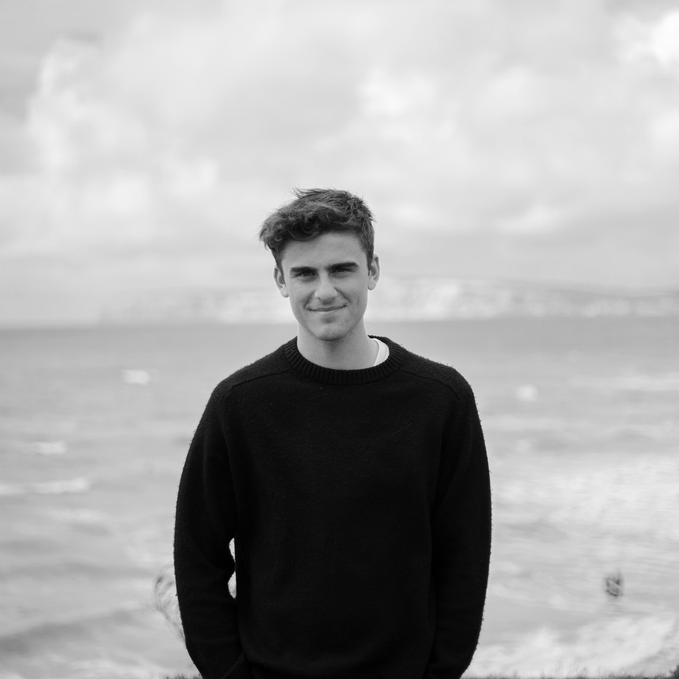 Black and white photograph of a young man standing outdoors near water, wearing a dark sweater, with a cloudy sky in the background.