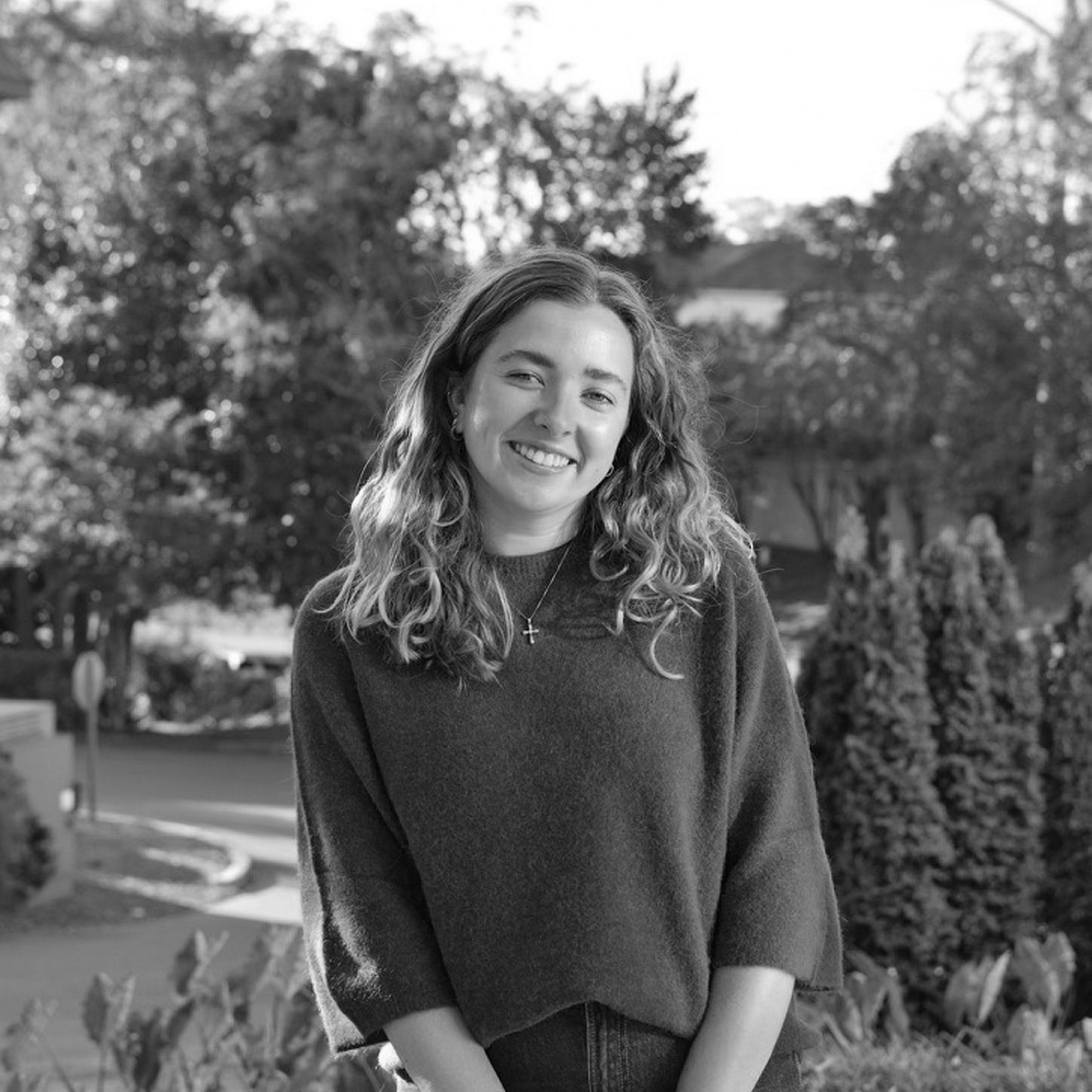 A smiling young woman with curly hair standing outdoors in a park, wearing a dark sweater and a cross necklace.