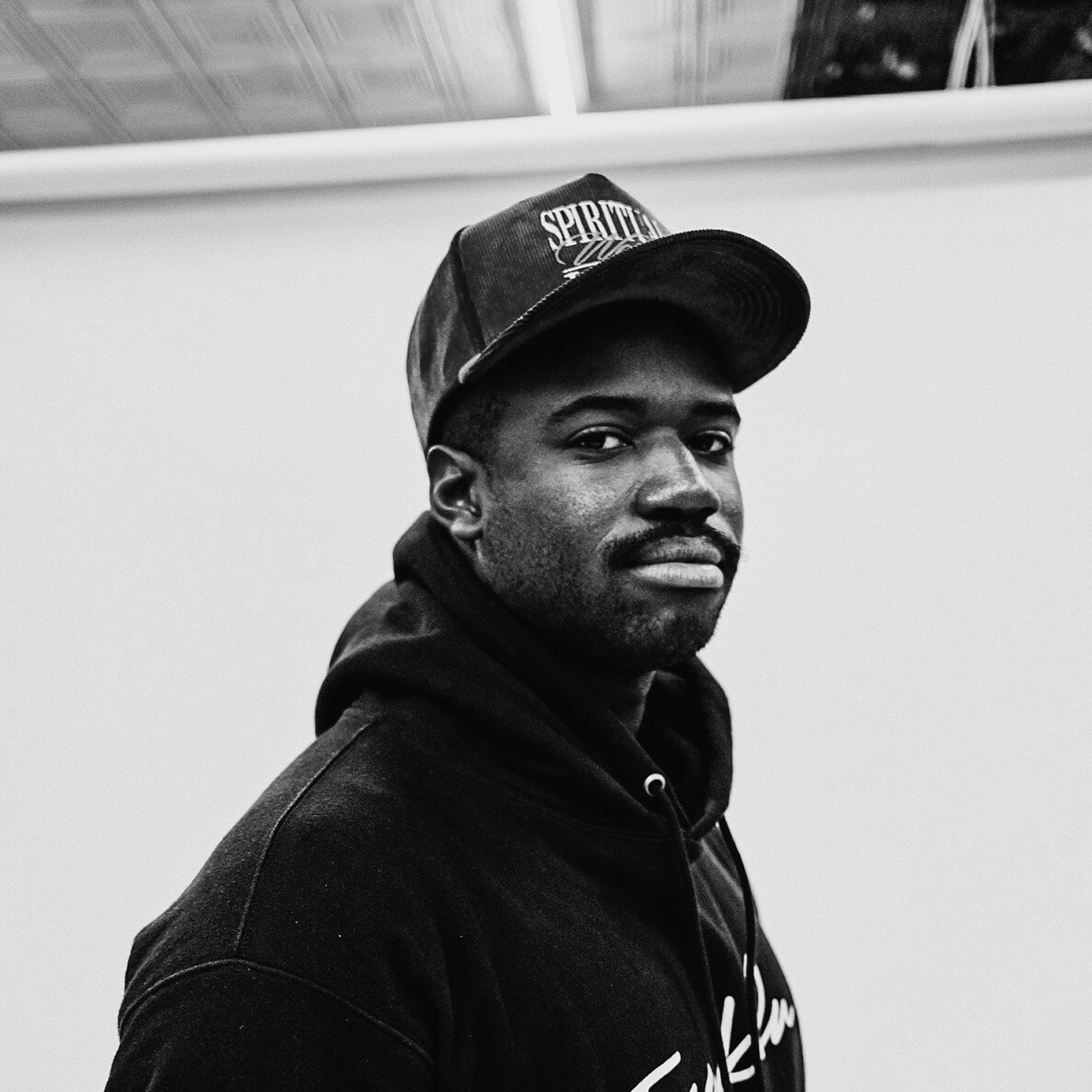 Black and white photo of a young man wearing a hoodie and a baseball cap, looking at the camera with a neutral expression, against a plain background.
