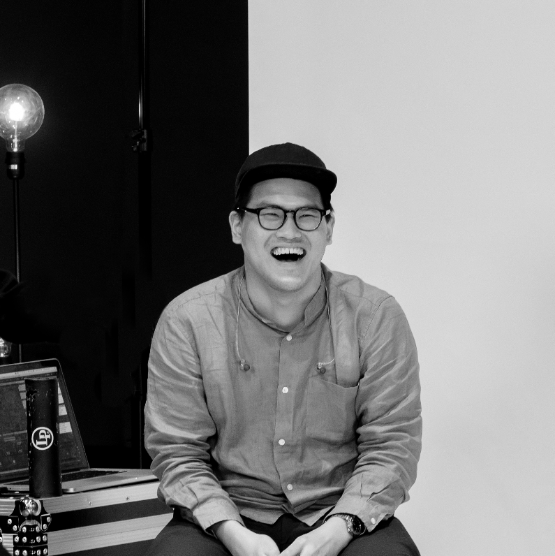A young man with glasses and a cap, laughing while sitting, with a person pointing at him from the left side of the image, in a room with a laptop and a light bulb in the background.