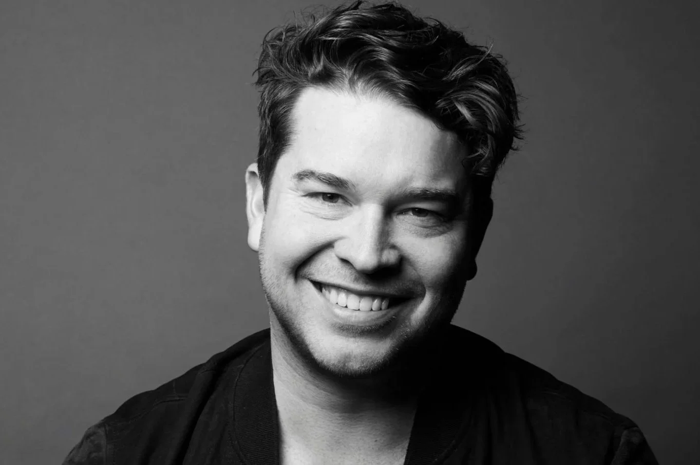 Black and white portrait of a smiling man with tousled hair, wearing a dark jacket, against a plain background.
