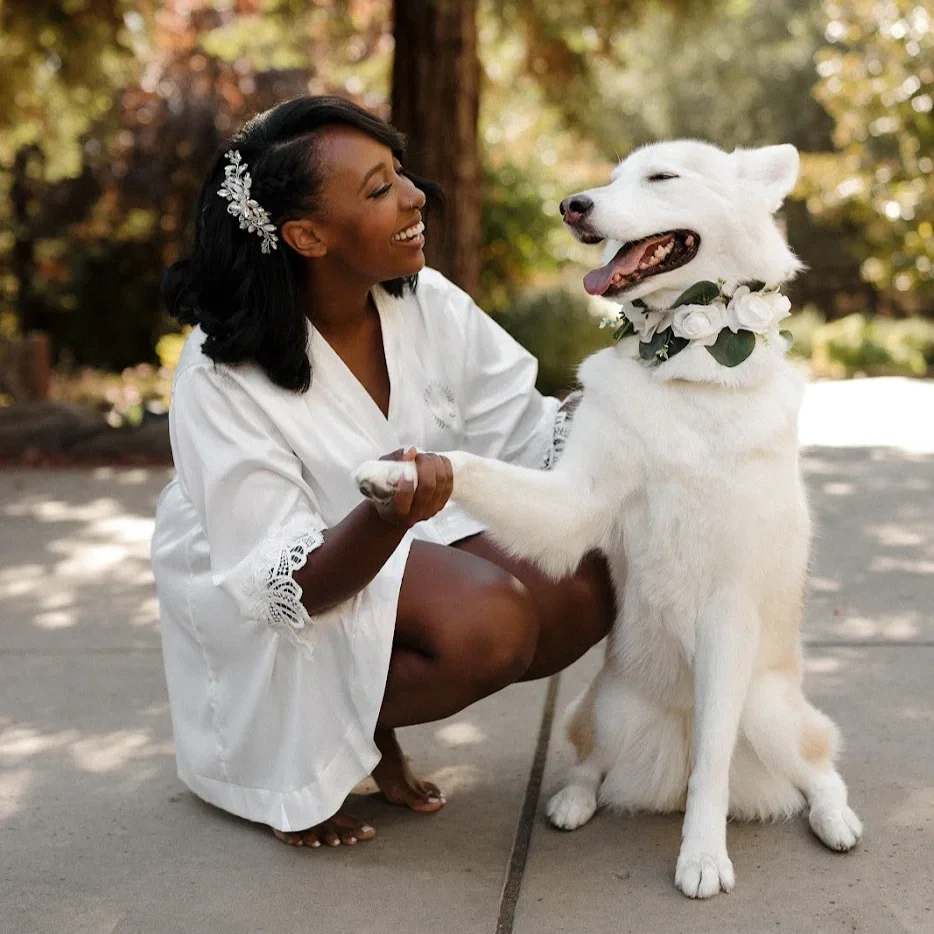 A bride kneels on the ground, holding hands with her Husky German Shepher mix dog wearing a floral collar. The bride is smiling and looking at the dog, and the dog appears happy with its tongue out. They are outdoors with trees in the background.