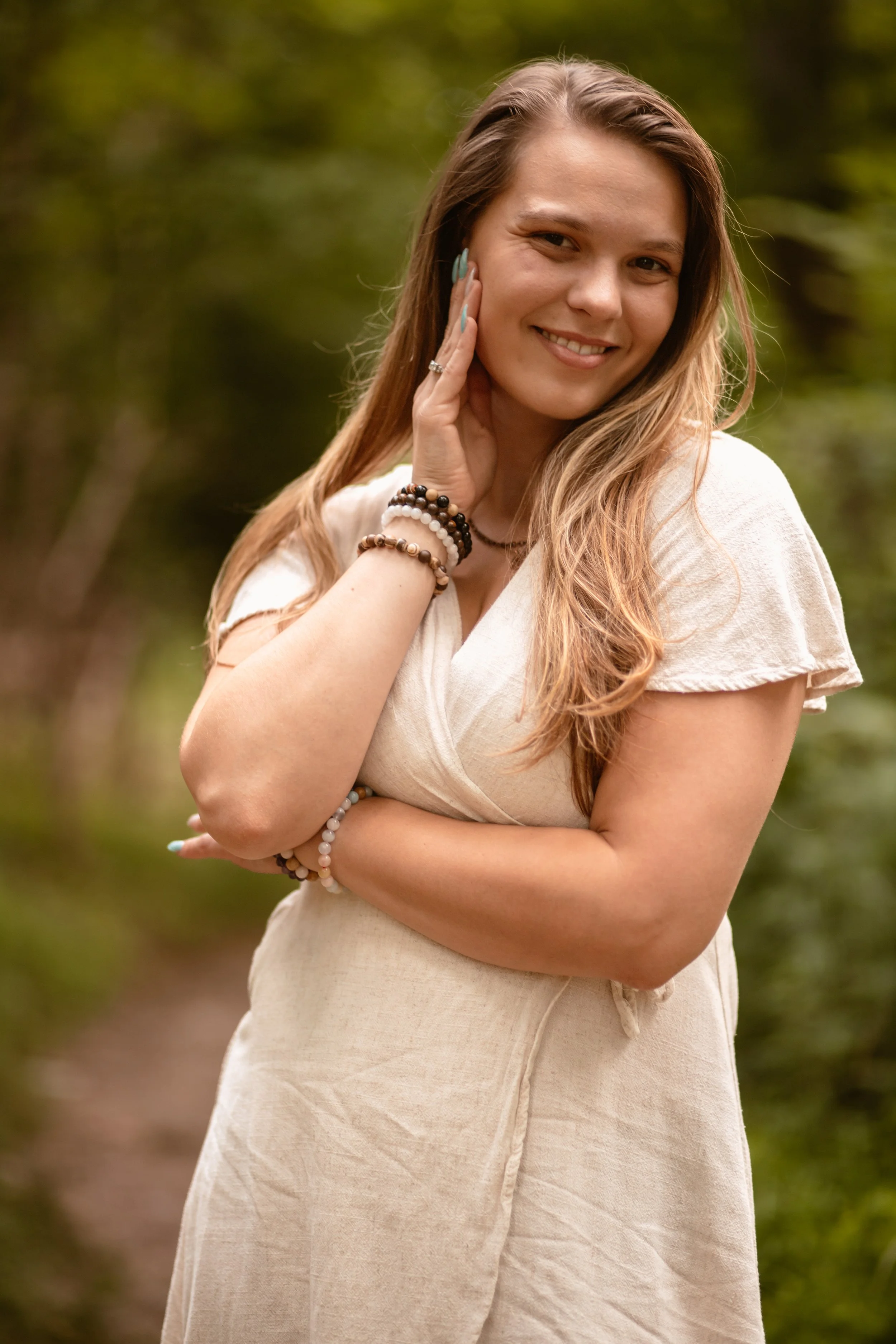 A smiling woman with long, wavy brown hair stands outdoors on a nature trail, wearing a beige dress and multiple bracelets, with her hand touching her face.