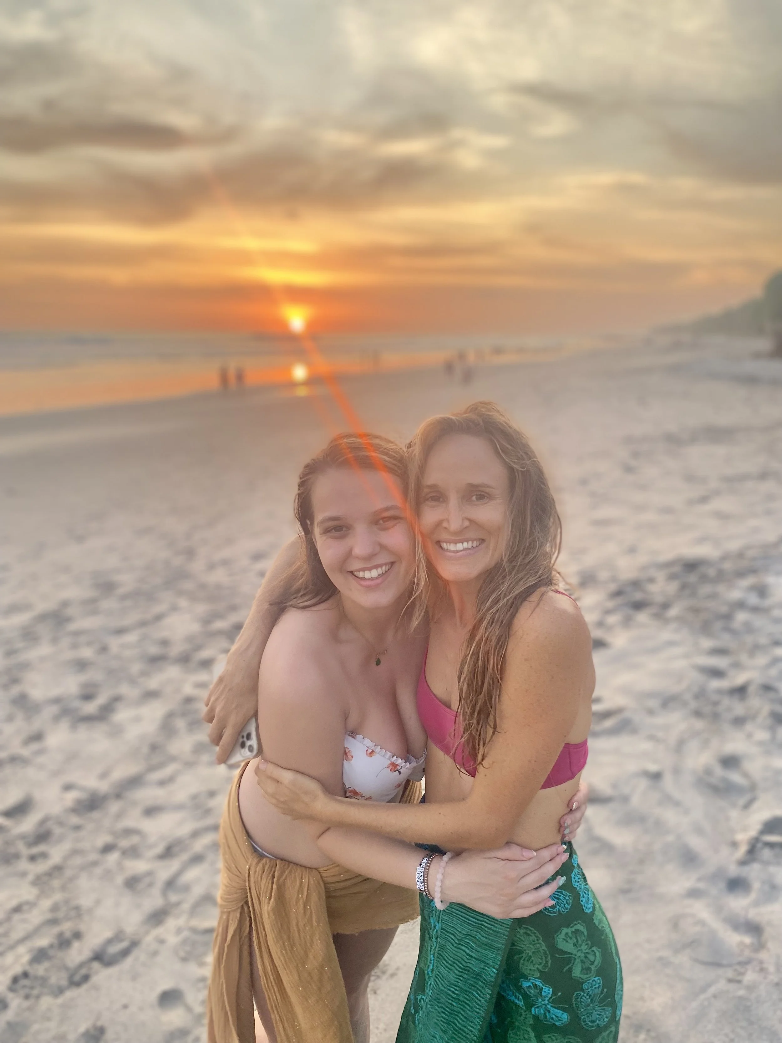 Two women hugging on the beach during sunset, smiling at the camera.