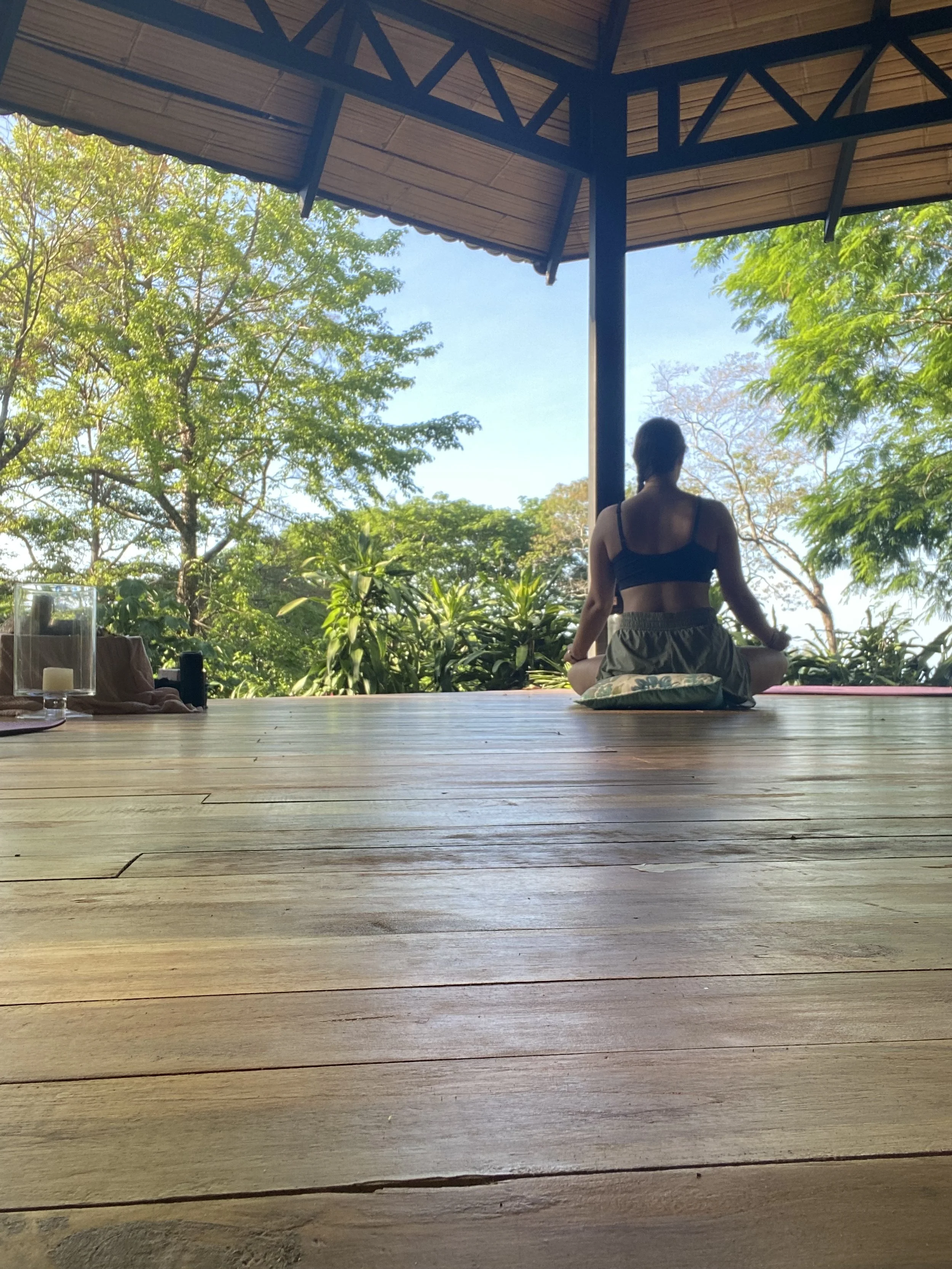 A person sitting cross-legged on a cushion meditating on a wooden deck, facing a lush green trees and blue sky view under a thatched-roof structure.