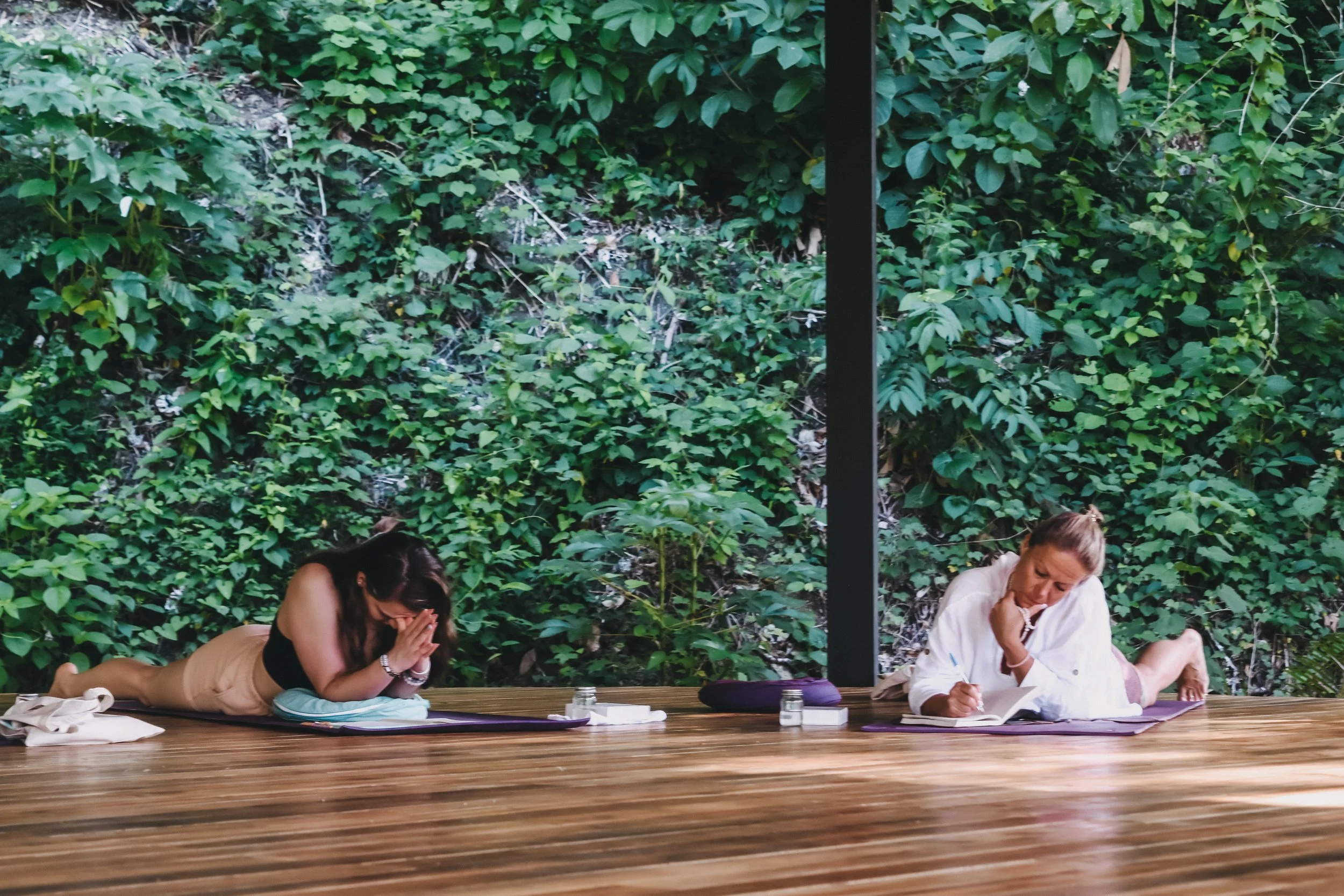 Two women practicing yoga outdoors on a wooden deck, surrounded by lush green foliage, with prayer or meditation poses.