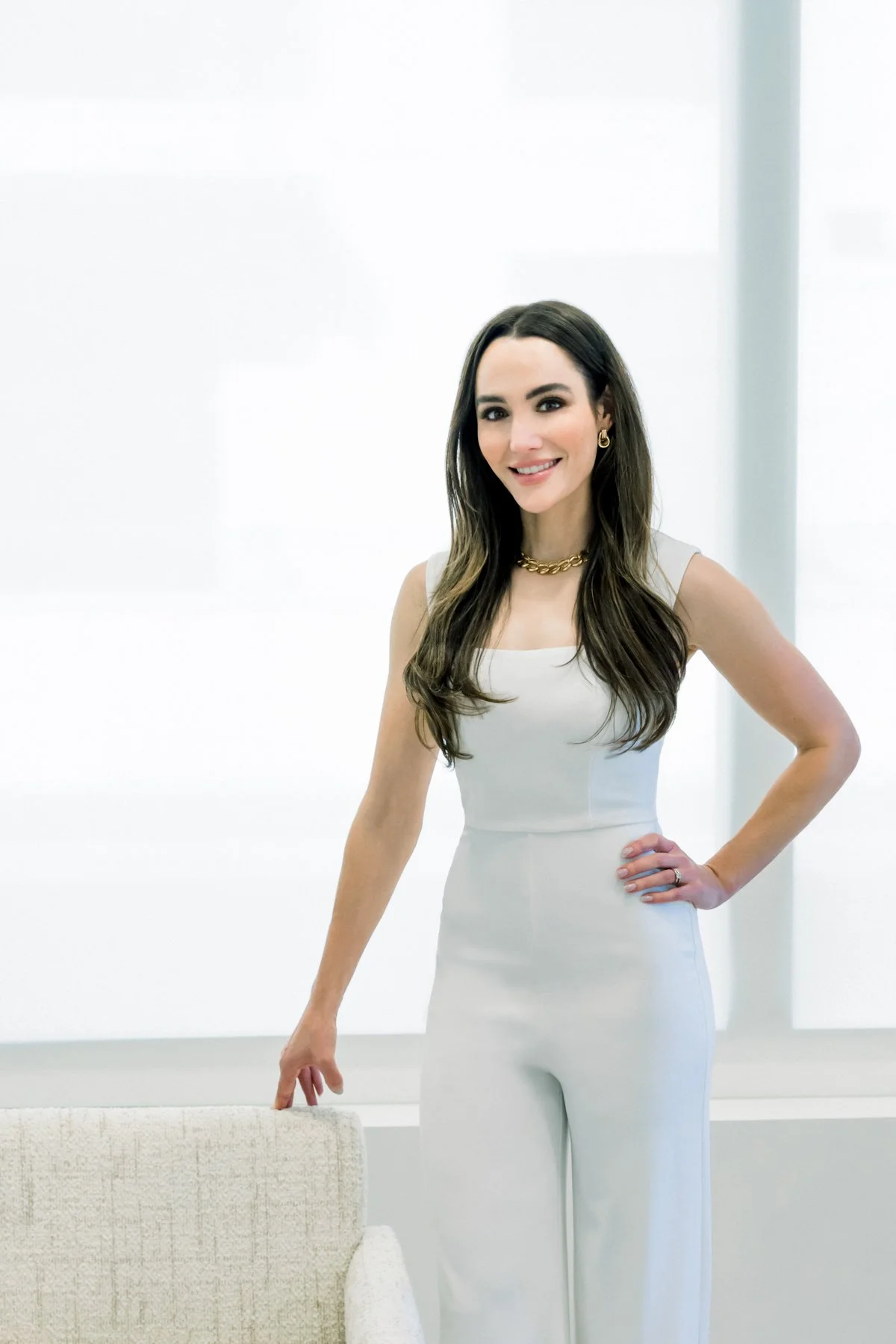 Woman in a white sleeveless jumpsuit standing indoors, smiling, with long brown hair, wearing gold jewelry, and standing next to a white chair.
