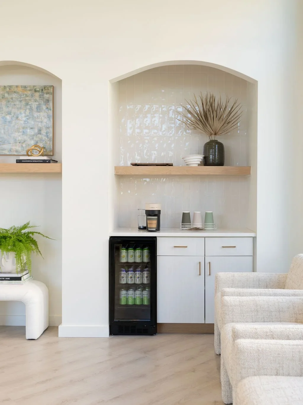 A small bar area with a mini refrigerator, coffee maker, and three cups, set in a white cabinet with gold handles beneath a wooden shelf, decorated with a black vase holding dried palm leaves and a white bowl on top.