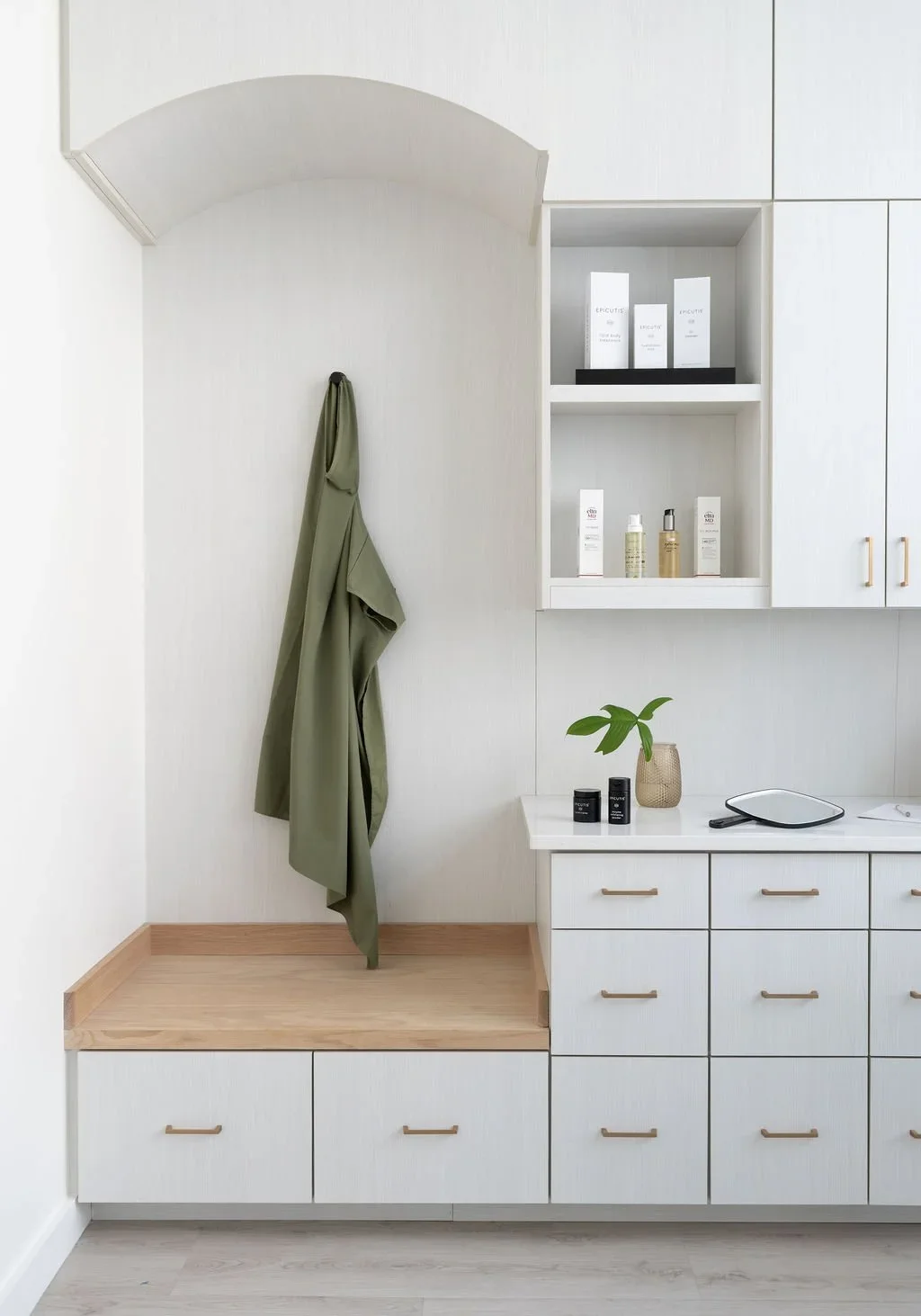 Minimalist white and light wood entryway with a coat hanging from a hook, a sideboard with a plant and toiletries, and wall-mounted cabinets with skincare products.