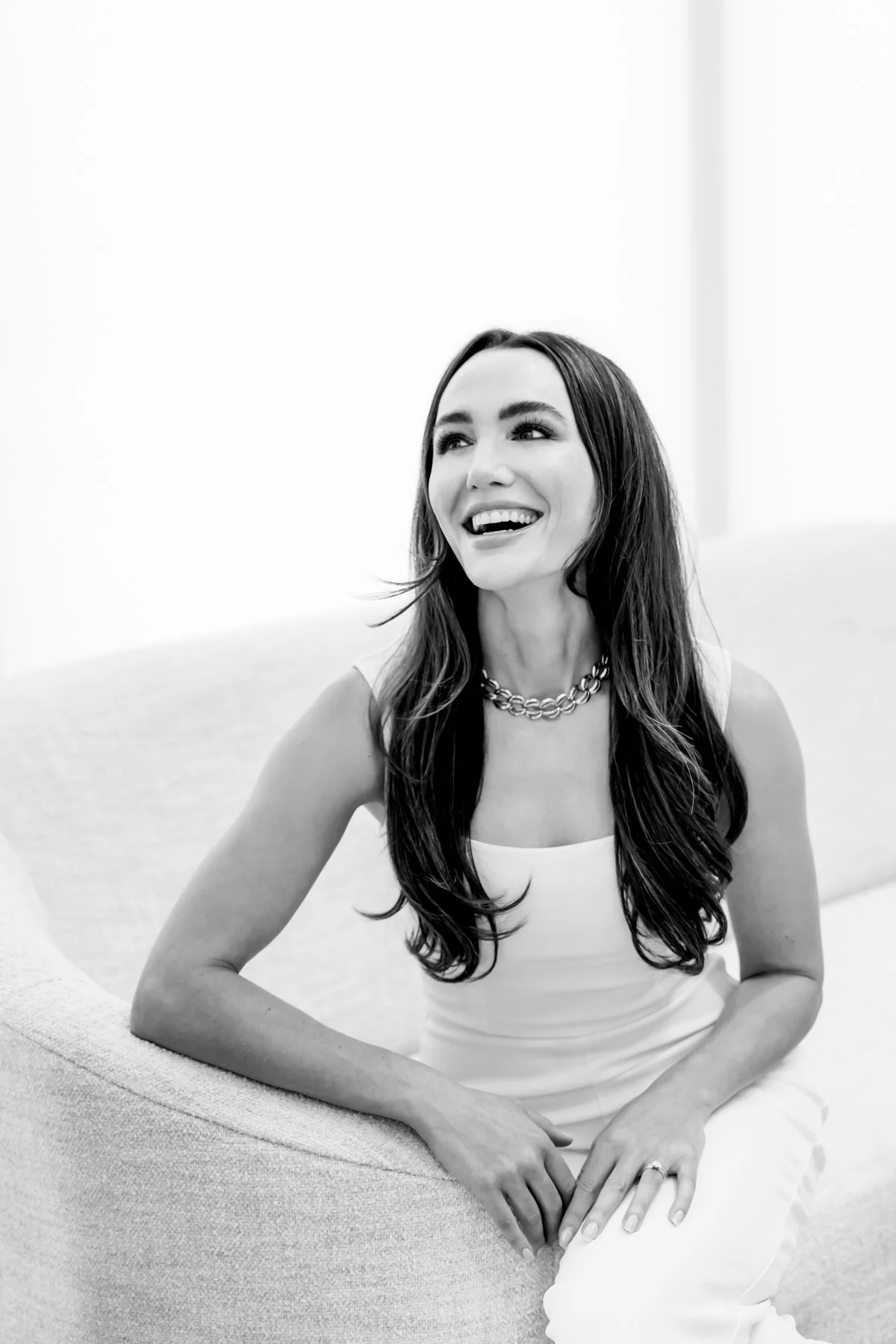 A woman with long dark hair and a broad smile sitting on a light-colored sofa, wearing a sleeveless top, a chain necklace, and a ring on her finger, in a minimalistic, bright room.