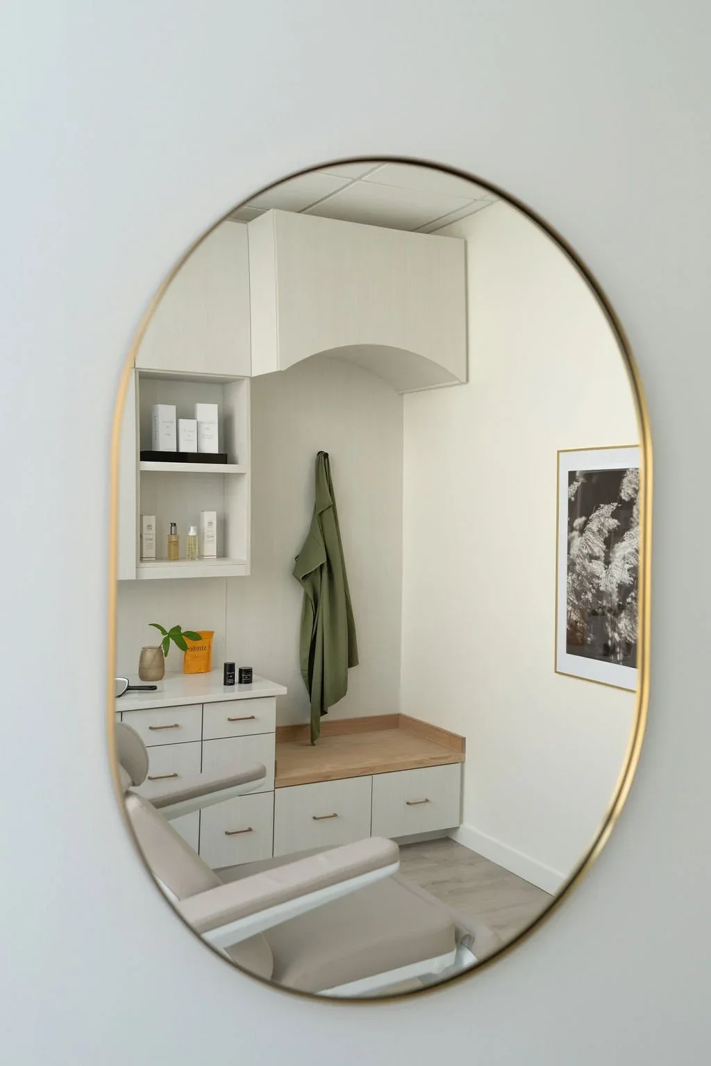 A reflection of a room in an oval mirror showing a white dresser, a beige chair, a green robe hanging on the wall, a framed black and white floral picture, and shelves with skincare products.