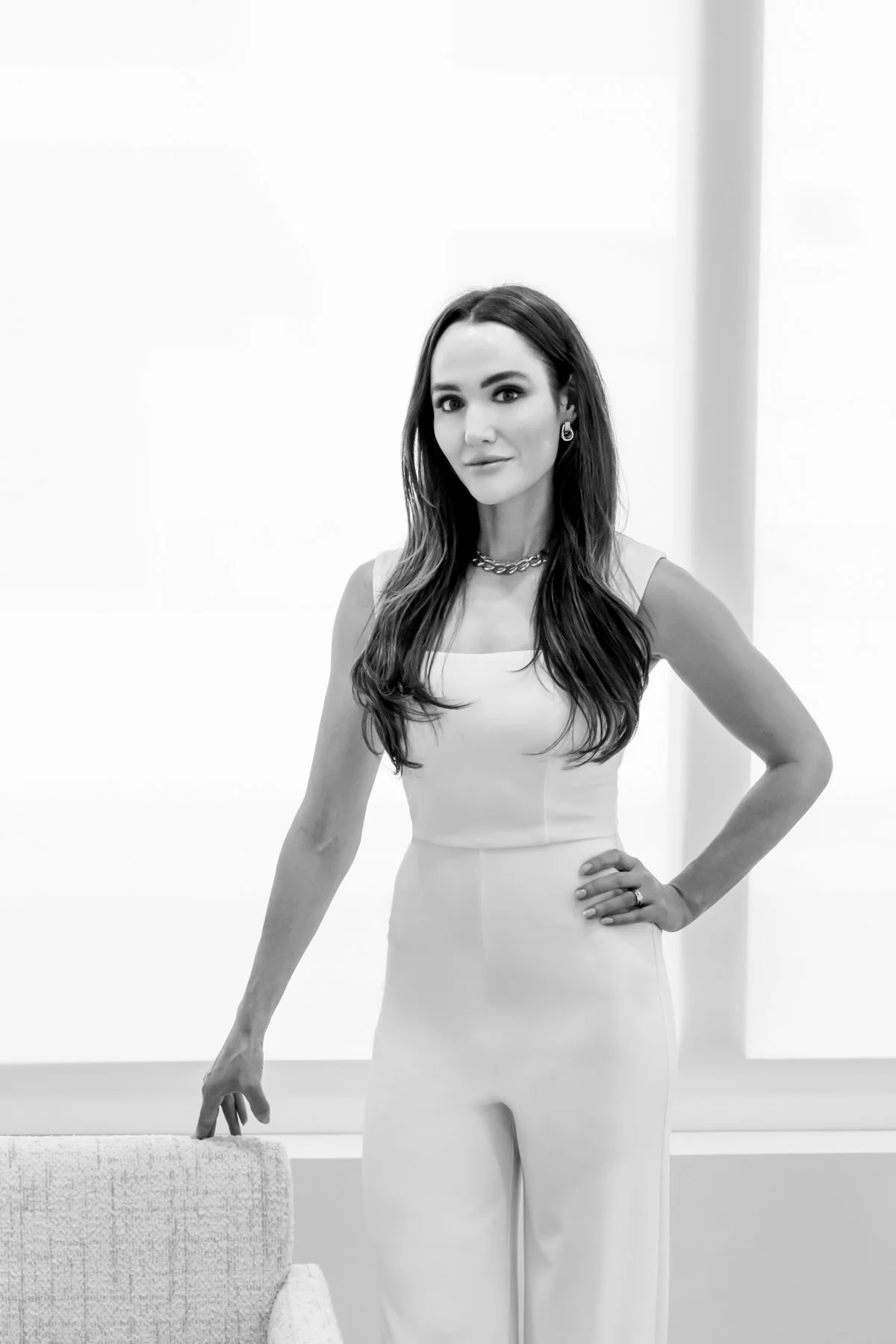 Black and white photo of a woman in a sleeveless dress, standing with one hand on her hip and the other resting on a chair, indoors with bright lighting.