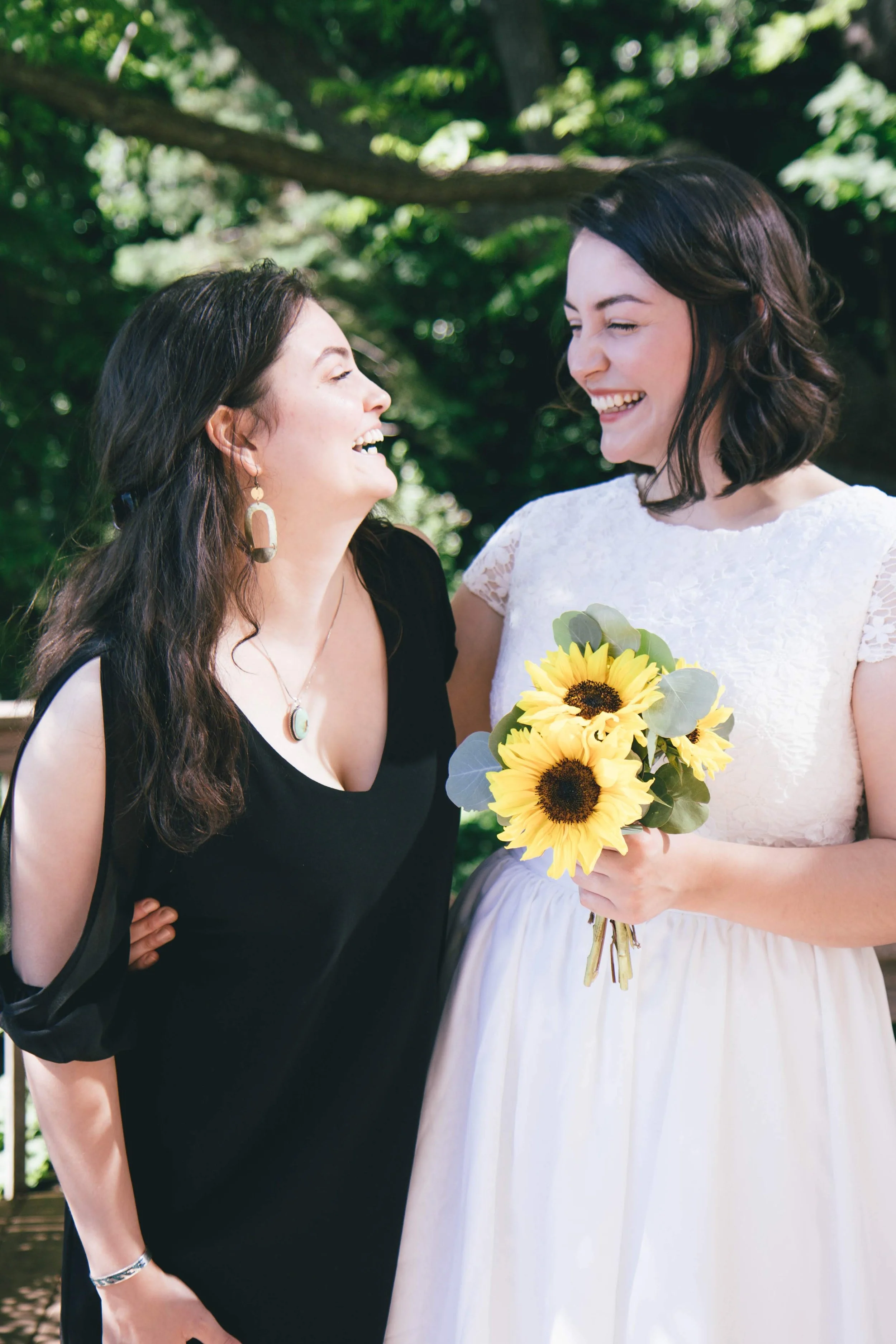 Two women smiling and laughing at a wedding, one wearing a black dress and the other wearing a white wedding dress, holding a bouquet of sunflowers. They are outdoors with lush green tree foliage in the background.