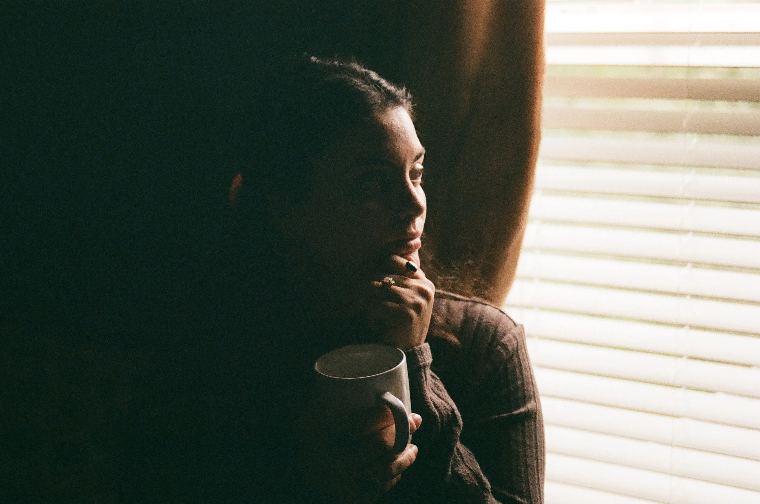 A woman sitting indoors near window blinds, holding a mug, with sunlight illuminating her face.