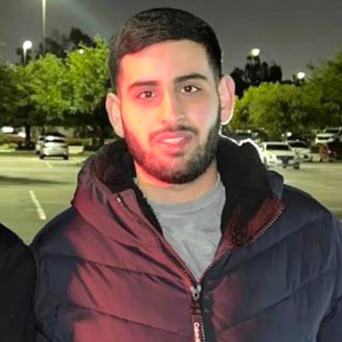 A young man with a beard and short dark hair, wearing a black and burgundy jacket and gray shirt, standing in a parking lot at night with trees and parked cars in the background.