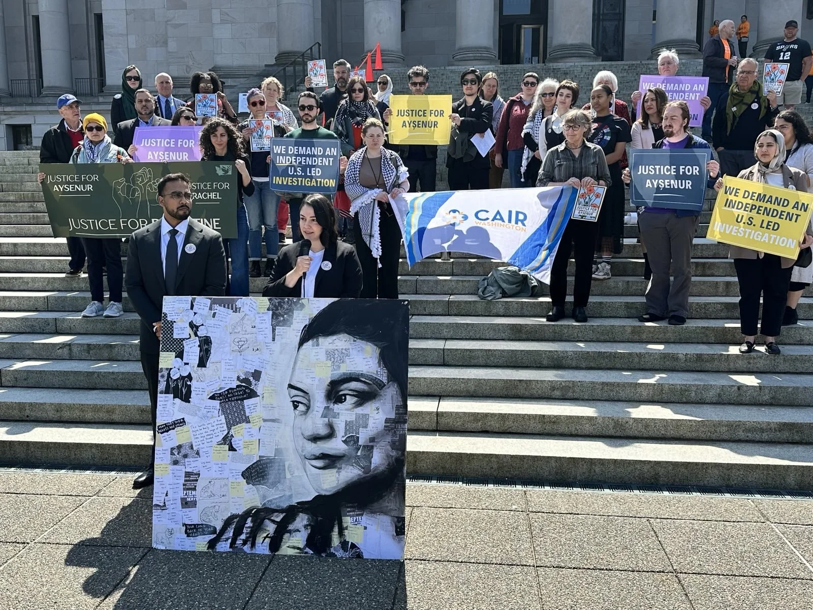 A group of people gathered on the stairs of a government building holding signs and banners advocating for justice, including signs reading "Justice for Aysenur," "Justice for Rachel," and "We demand an independent U.S. led investigation." There is an artist's portrait poster of a woman with notes and papers attached to it, placed in the foreground.