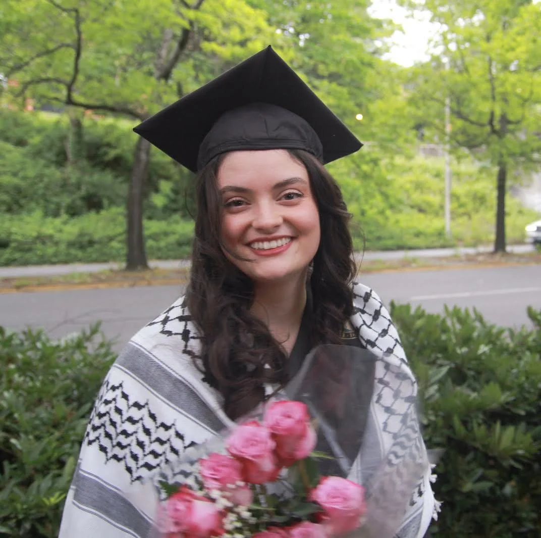 A young woman in a graduation cap and gown holding a bouquet of pink flowers outdoors with green trees and a street in the background.