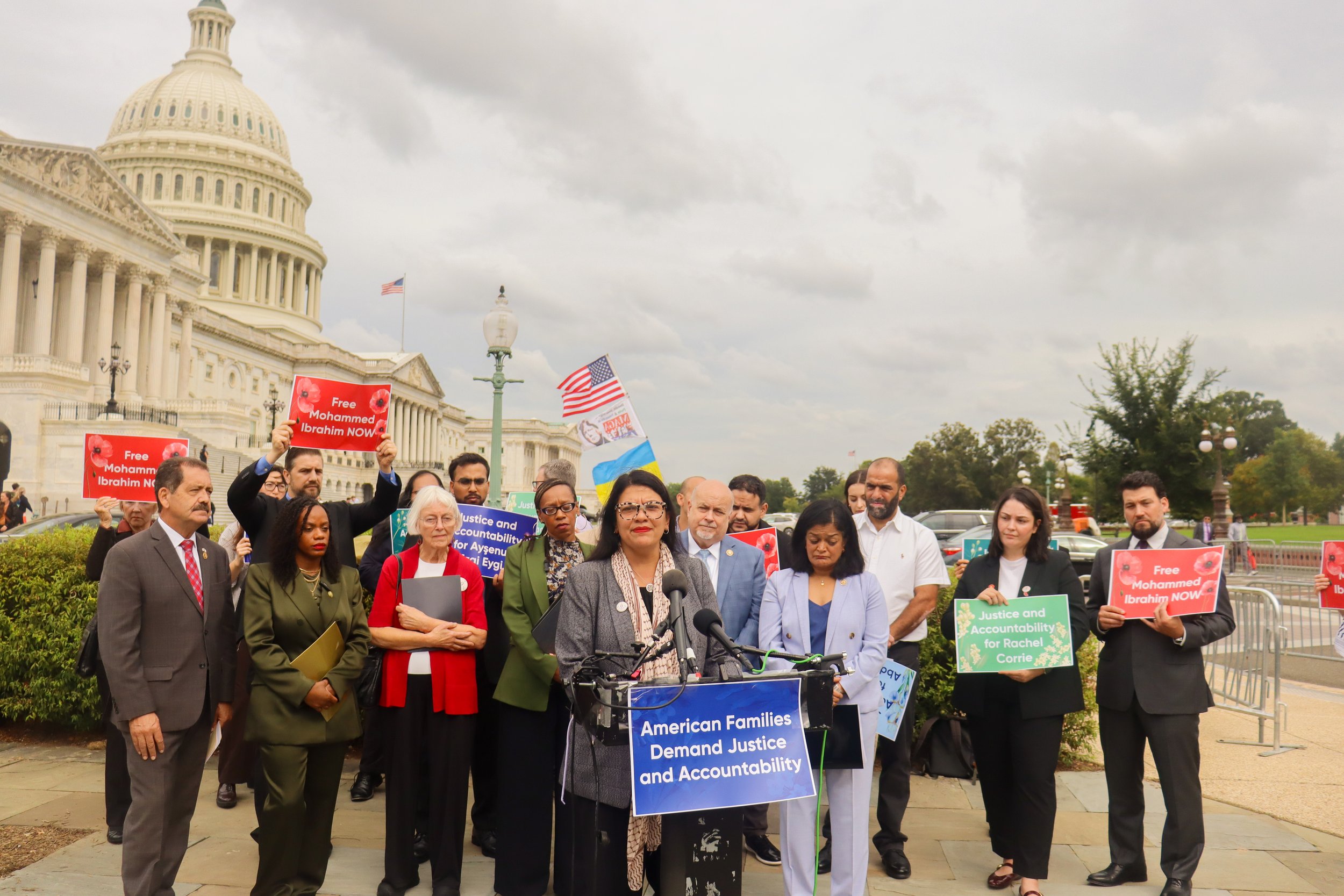 A diverse group of people participating in a protest or rally outside the U.S. Capitol building. They are holding signs advocating for justice and accountability for various causes, including freeing Mohamed Ibrahim and for Rachel Corrie. The scene is overcast with cloudy skies.