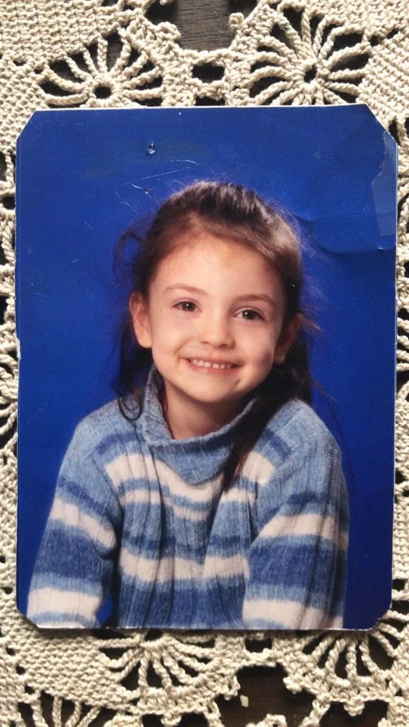 A young girl with brown hair pulled back, smiling, wearing a blue and white striped sweater, photo mounted on a textured lace tablecloth.