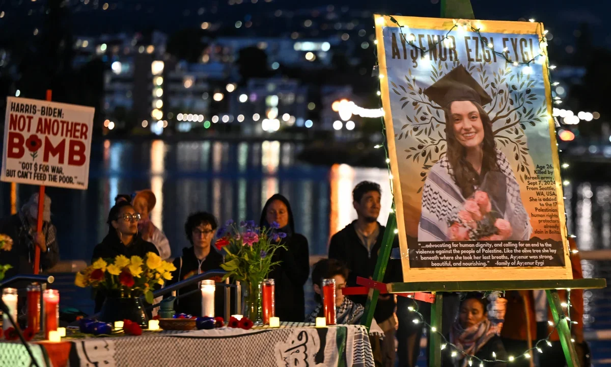 A memorial gathering at night by a waterfront with people holding signs, including a large portrait of a young woman in graduation cap and gown with flower bouquet, surrounded by flowers, candles, and lights, honoring Asaynur Eygi, who was killed in September 2020 from Seattle, WA, USA.