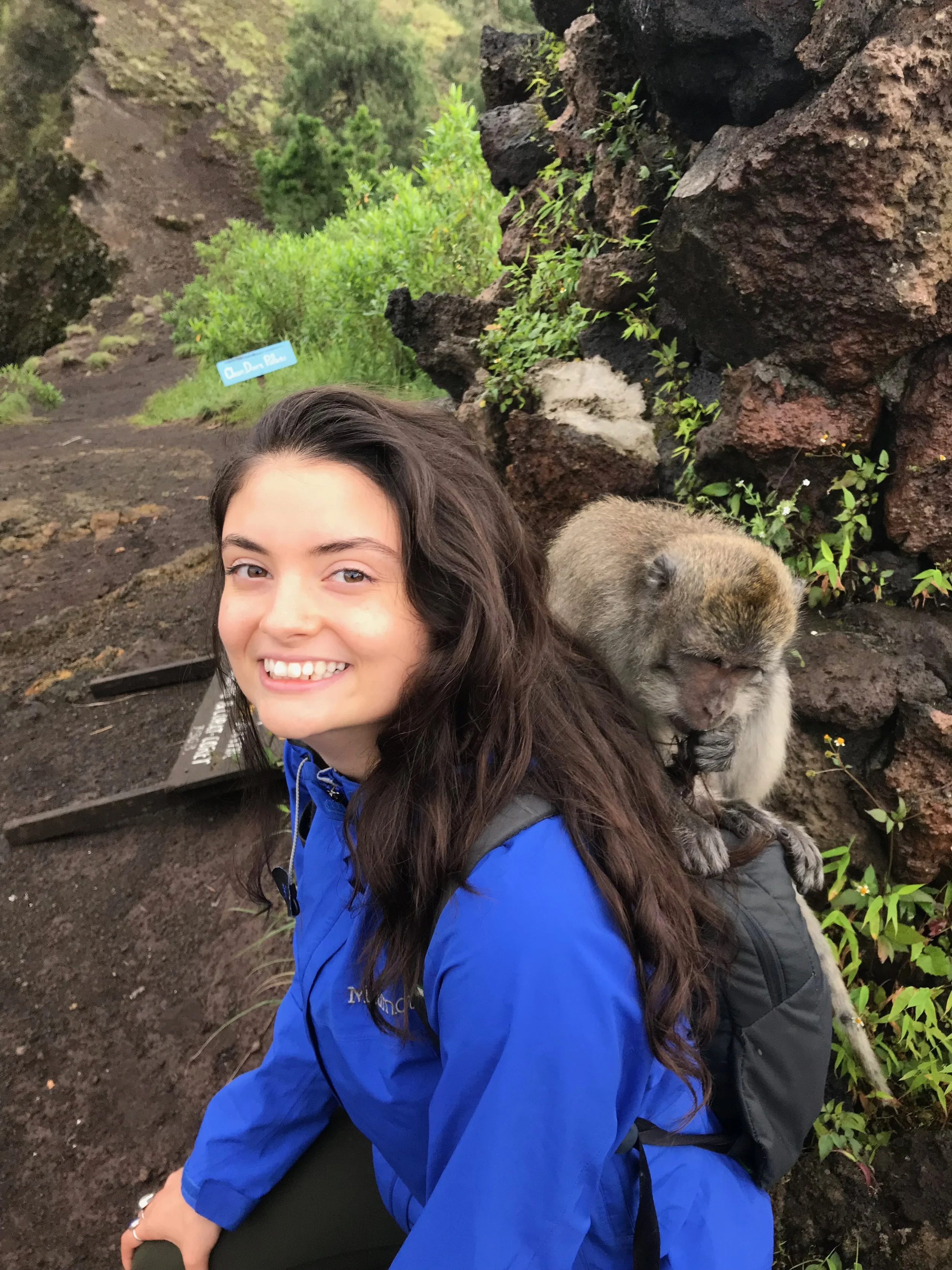 A smiling young woman with long dark hair, wearing a blue jacket, sitting on the ground with an adorable puppy, in front of a rocky, green, outdoor landscape.