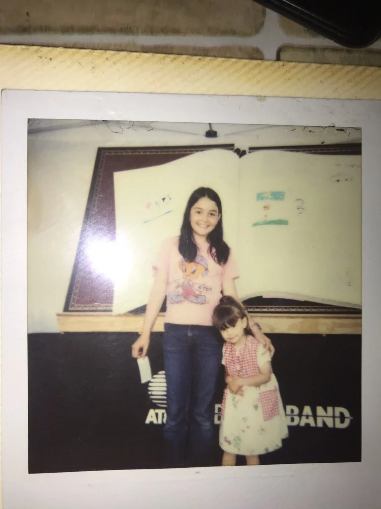 Two girls, one older and one younger, standing together in front of a blackboard with text that appears to say "AT THE BAND." The older girl is smiling, wearing a pink cartoon t-shirt and jeans, and holding a piece of paper. The younger girl is wearing a checkered top and a floral skirt, holding her hands together and leaning slightly against the older girl.