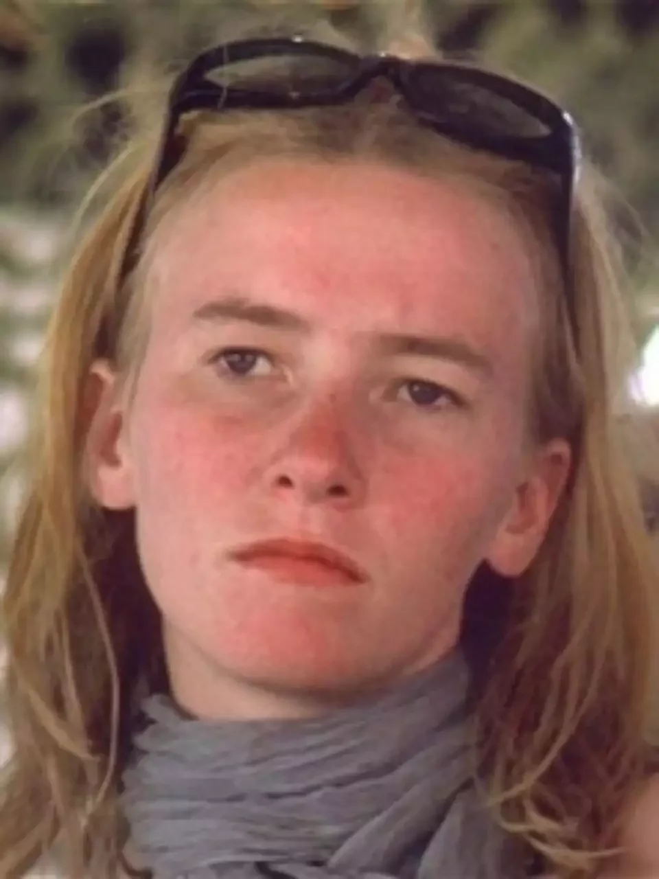Close-up of a young woman with long blonde hair, sunglasses on her head, wearing a gray scarf around her neck, with a serious expression on her face.