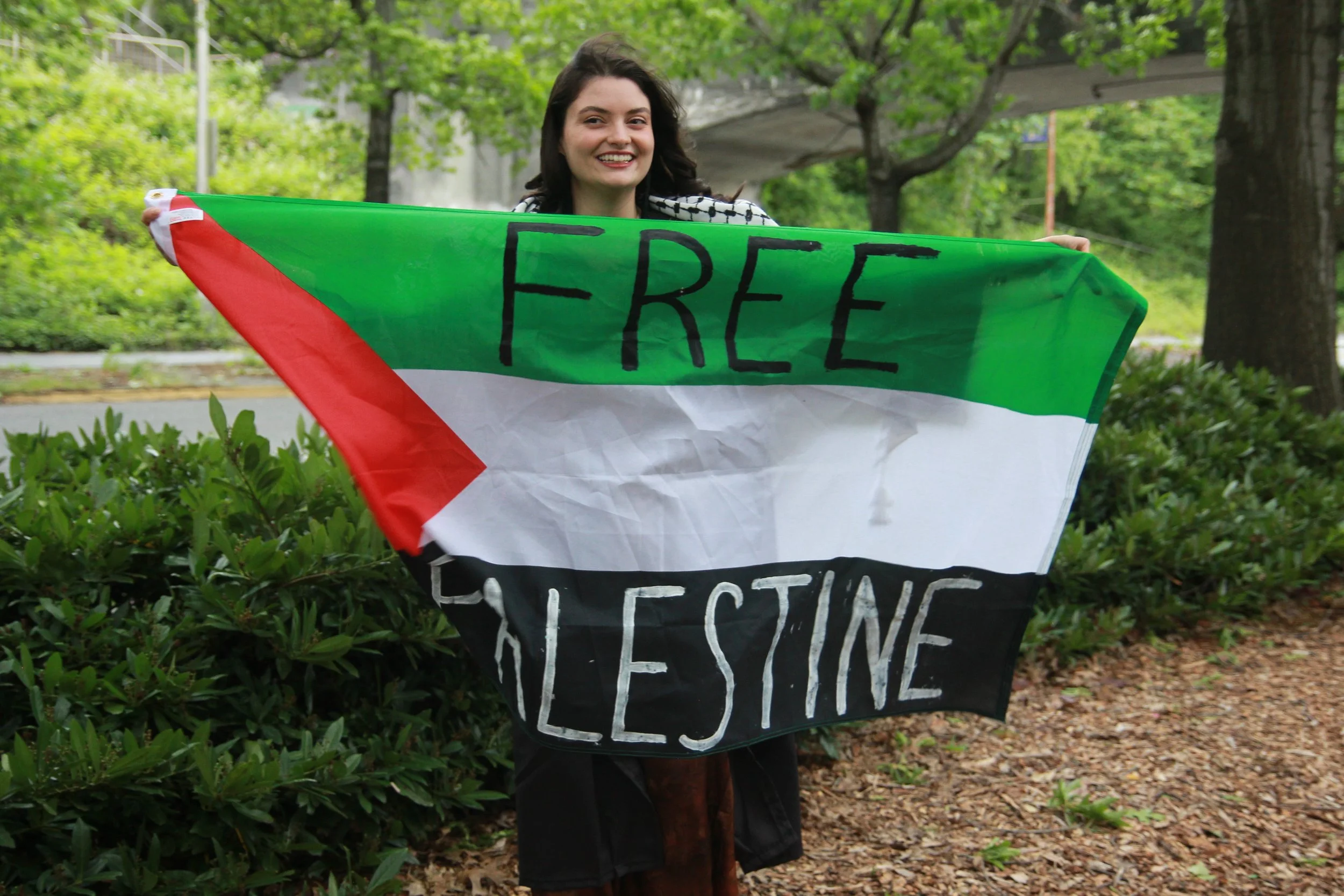 Woman holding a protest sign that reads 'FREE' with parts of other words visible, standing outdoors among trees and greenery.