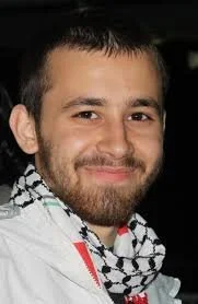 A smiling young man with short dark hair and a beard, wearing a white jacket and a black-and-white patterned scarf.